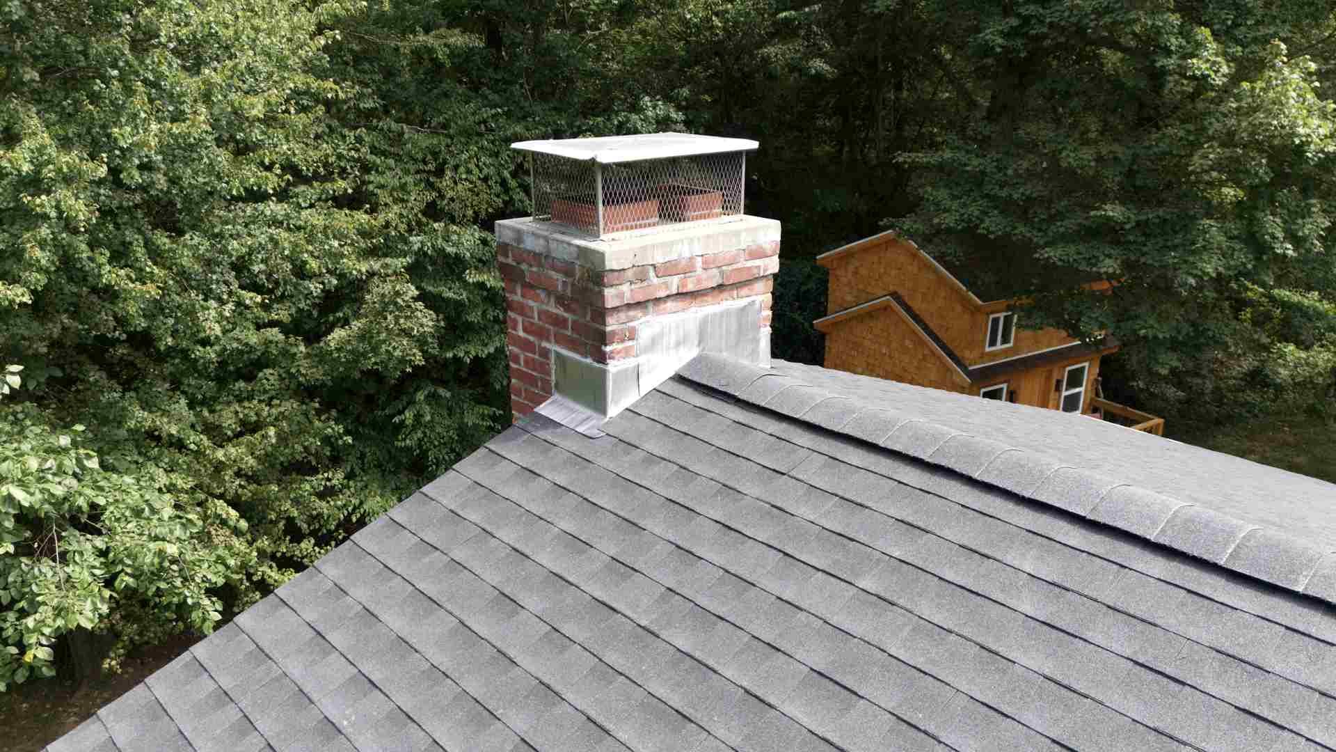 Gray shingled roof with a brick chimney, surrounded by green trees and a wooden deck in the background