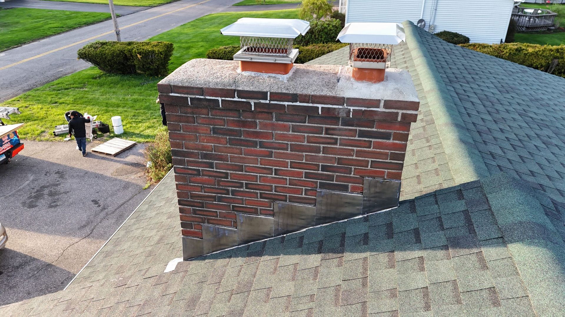Brick rooftop chimney with two metal caps on a shingled roof, seen from above.