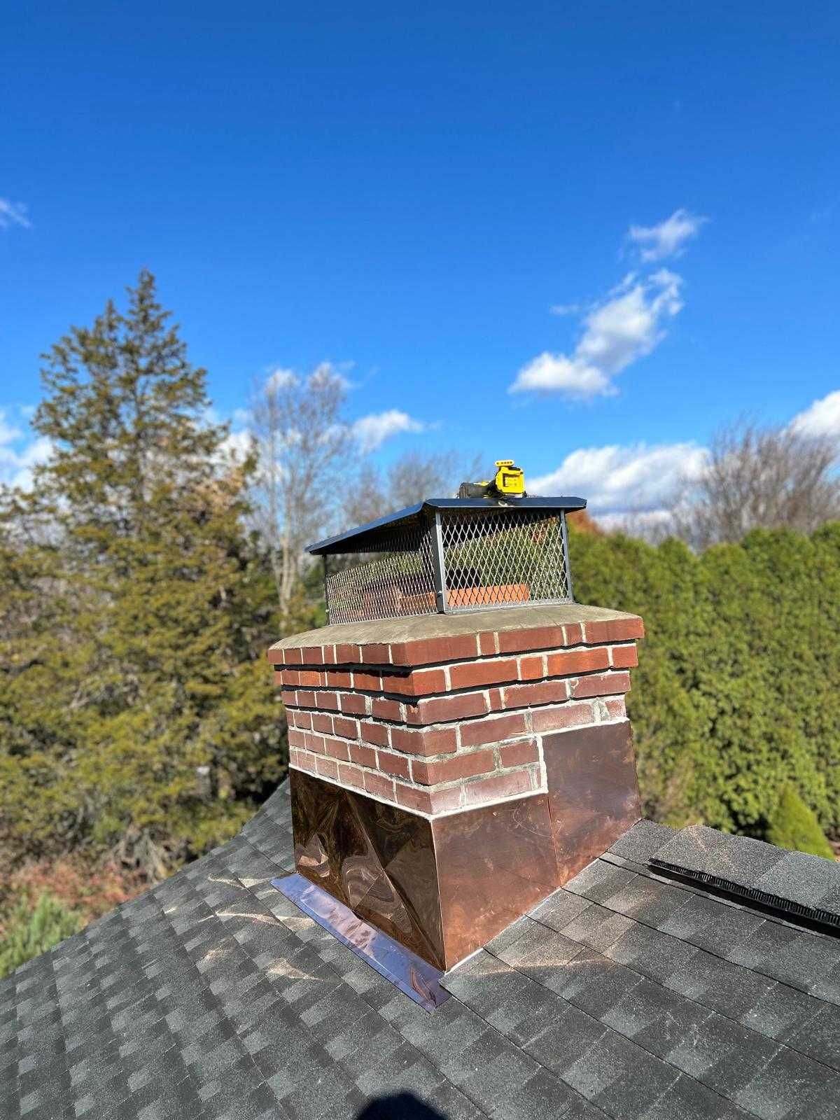 Brick chimney on a gray shingled roof with a yellow cap, set against blue sky and trees.
