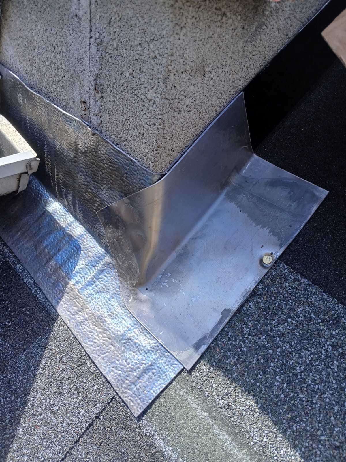 Metal flashing around a rooftop vent at the base of a concrete chimney on asphalt roofing.