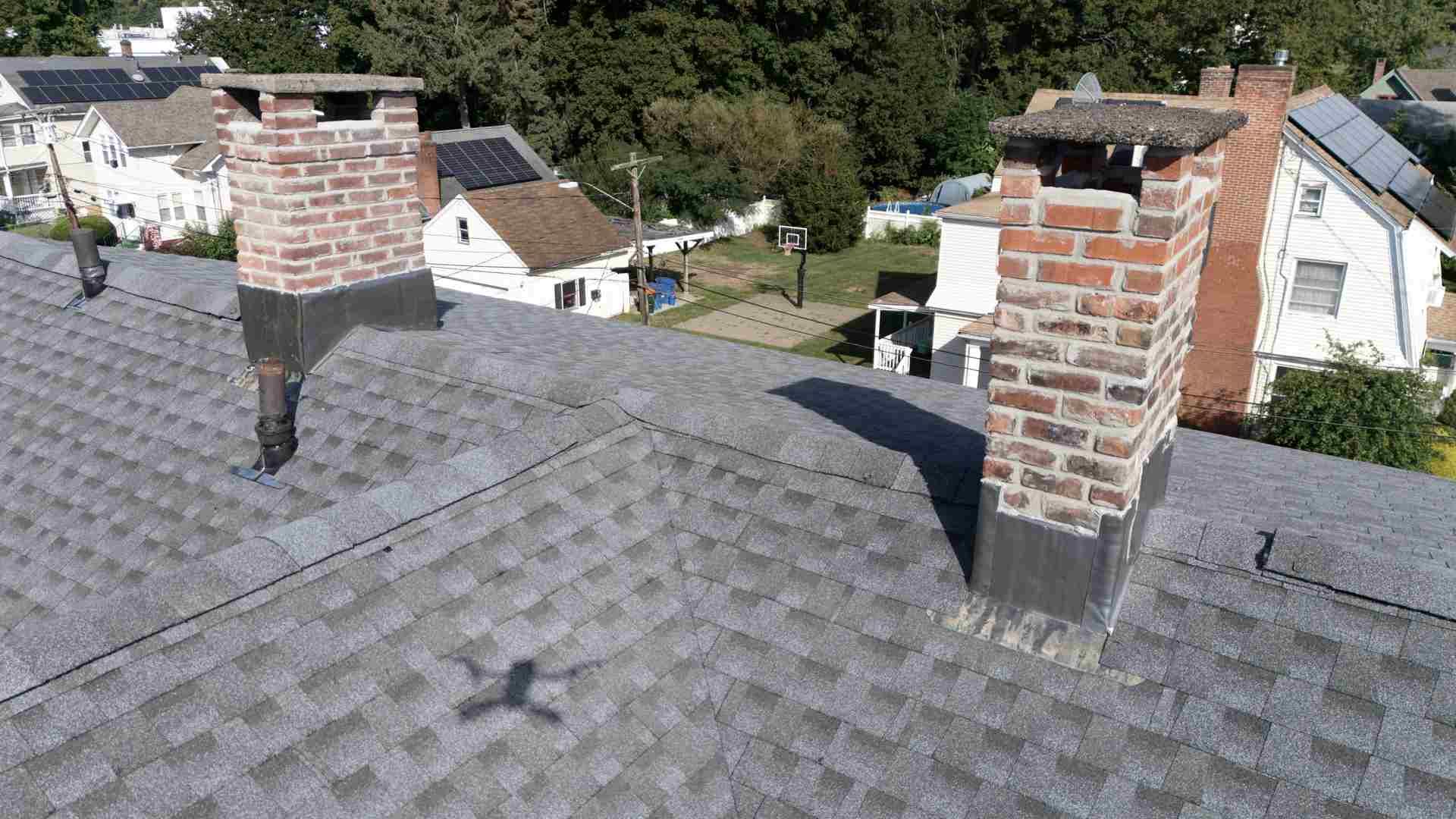 Rooftop with two brick chimneys on gray shingles, overlooking houses and trees.