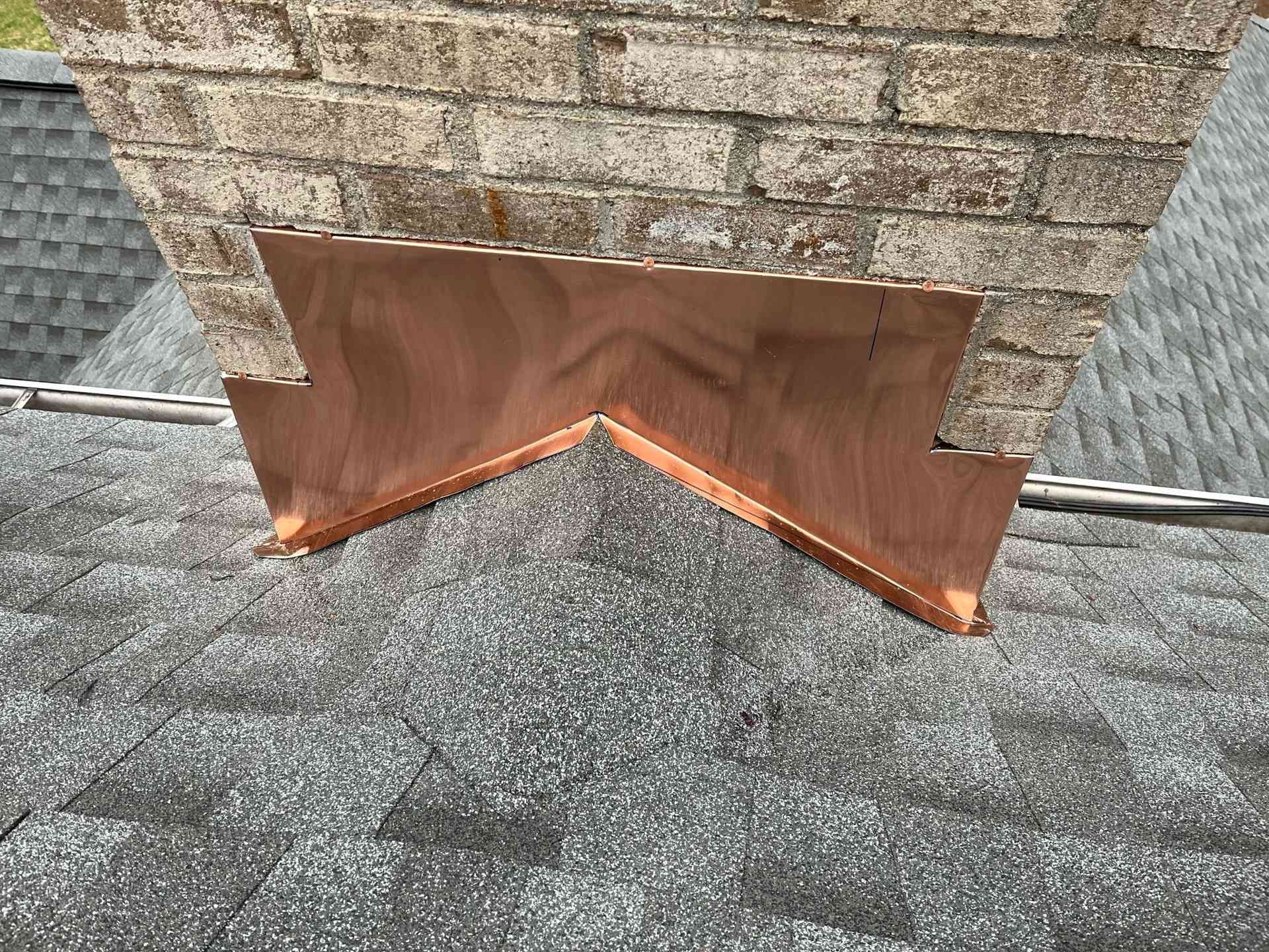 Copper roof flashing at the base of a brick chimney on a shingled roof.