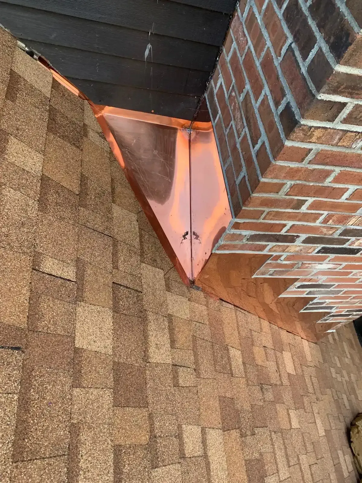 Roof valley with pink flashing between brown shingles and a brick wall