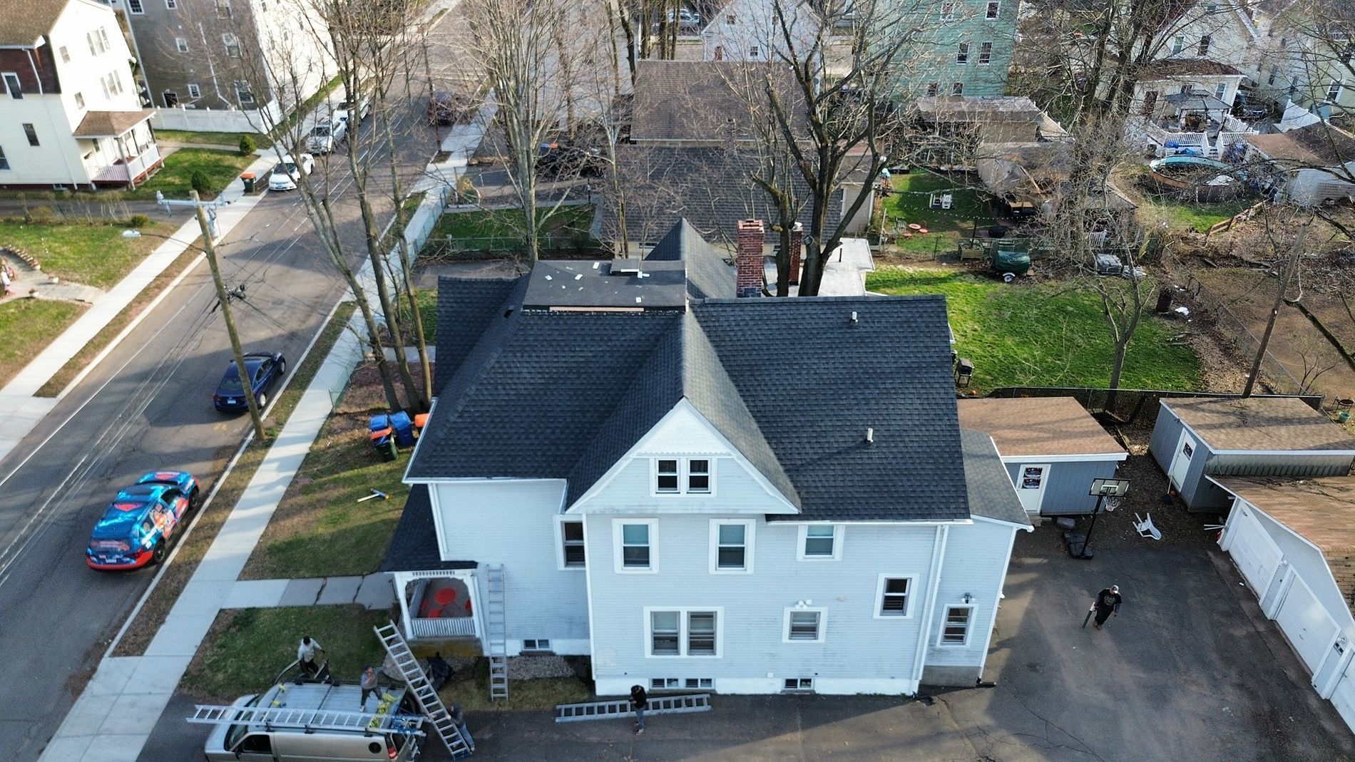 Aerial view of a white two-story house with black roof and parked cars in a suburban neighborhood.