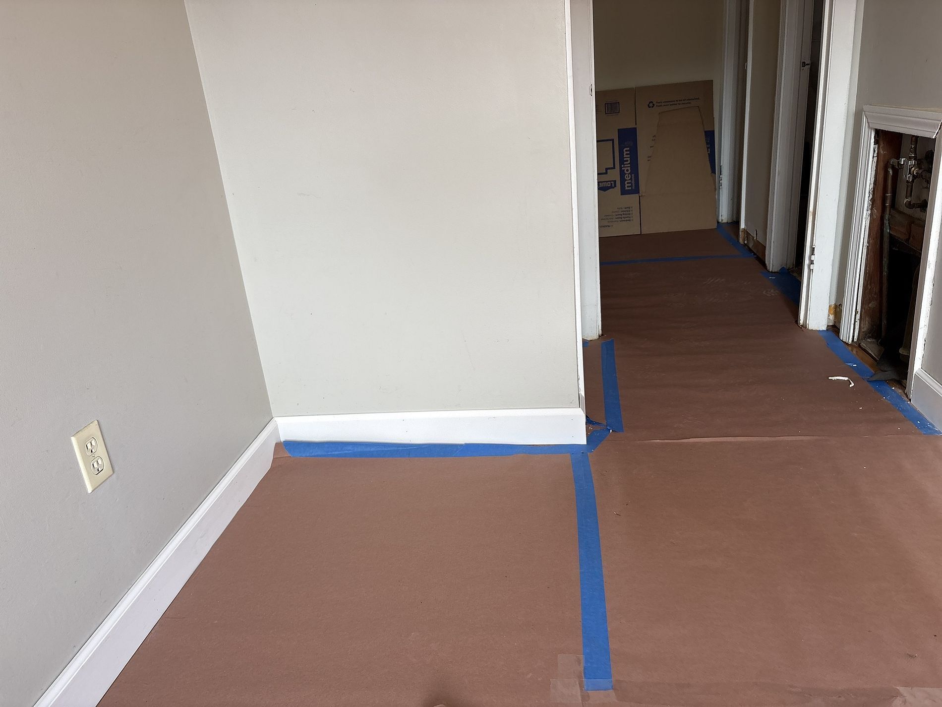 Hallway with brown floor covering taped down in blue, leading to a doorway and exposed wall opening.