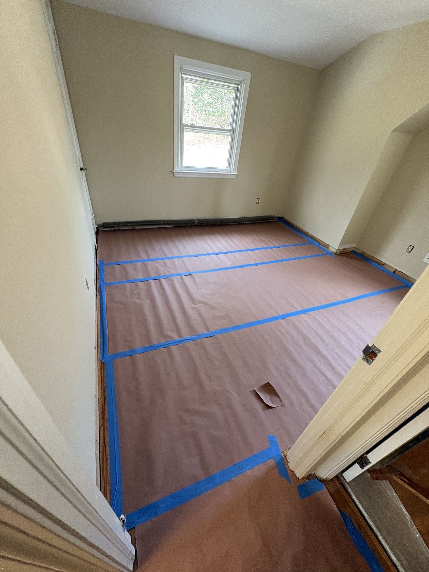 Small empty room with beige walls, single window, and brown floor covered in taped-down protective paper.
