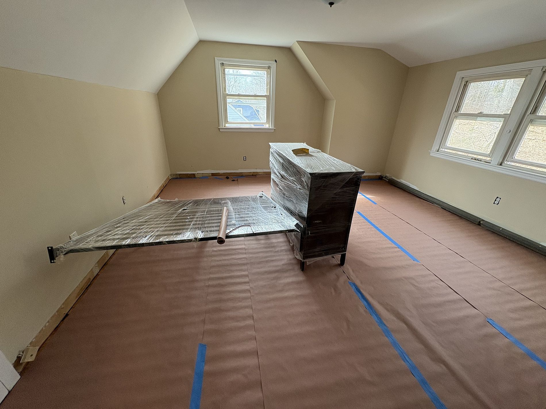 Empty attic room with sloped beige walls, brown floor, and a tarp-covered island in the center.