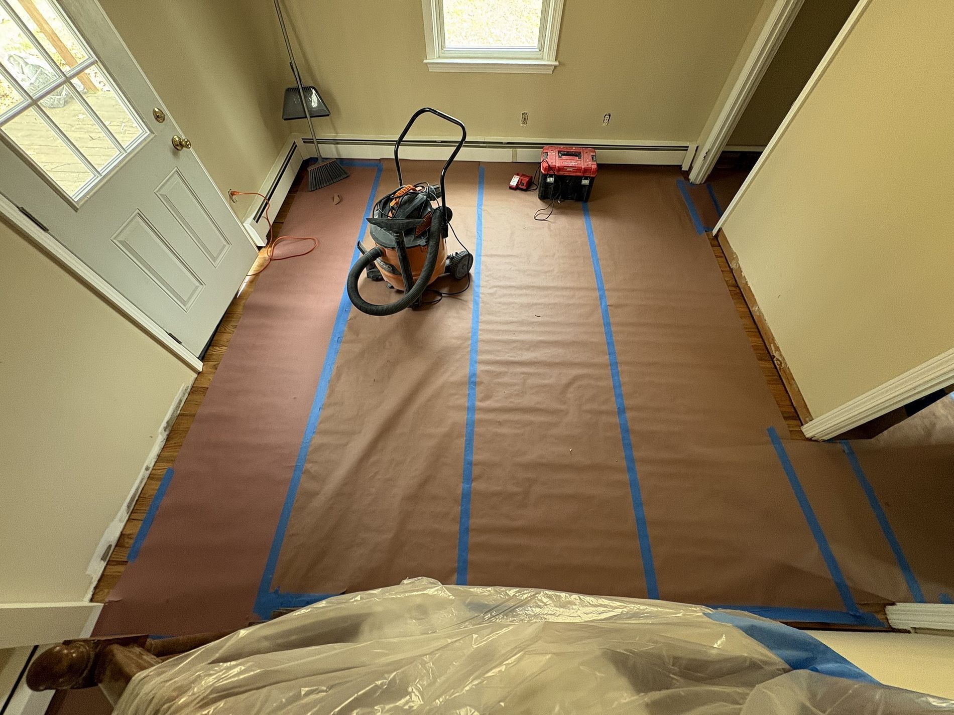 Room under renovation with plastic-covered floor, blue tape, a shop vacuum, and a red tool near a door and window