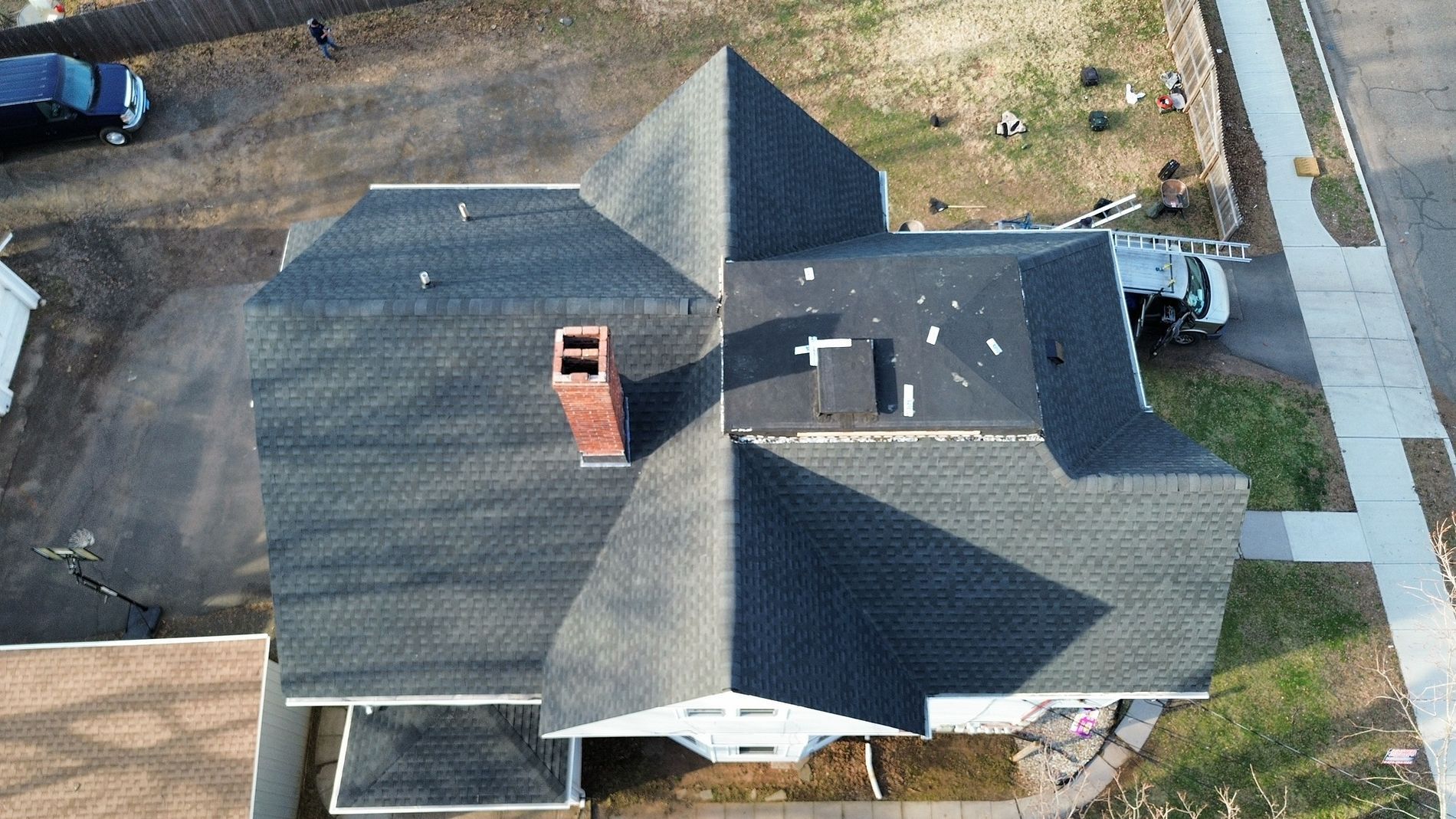 Aerial view of a house with a dark shingled roof, brick chimney, and driveway.