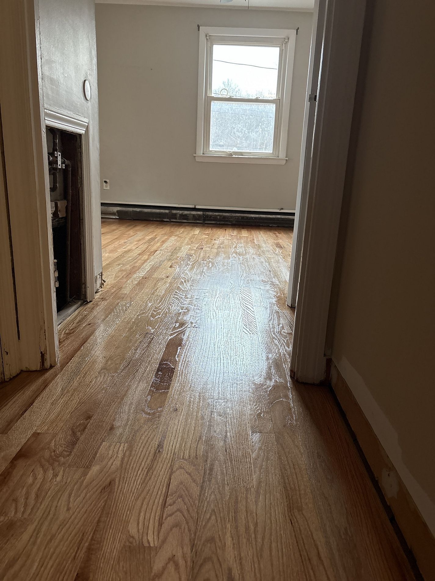 Narrow hallway with hardwood floor leading to a small room with a window and exposed fireplace opening.