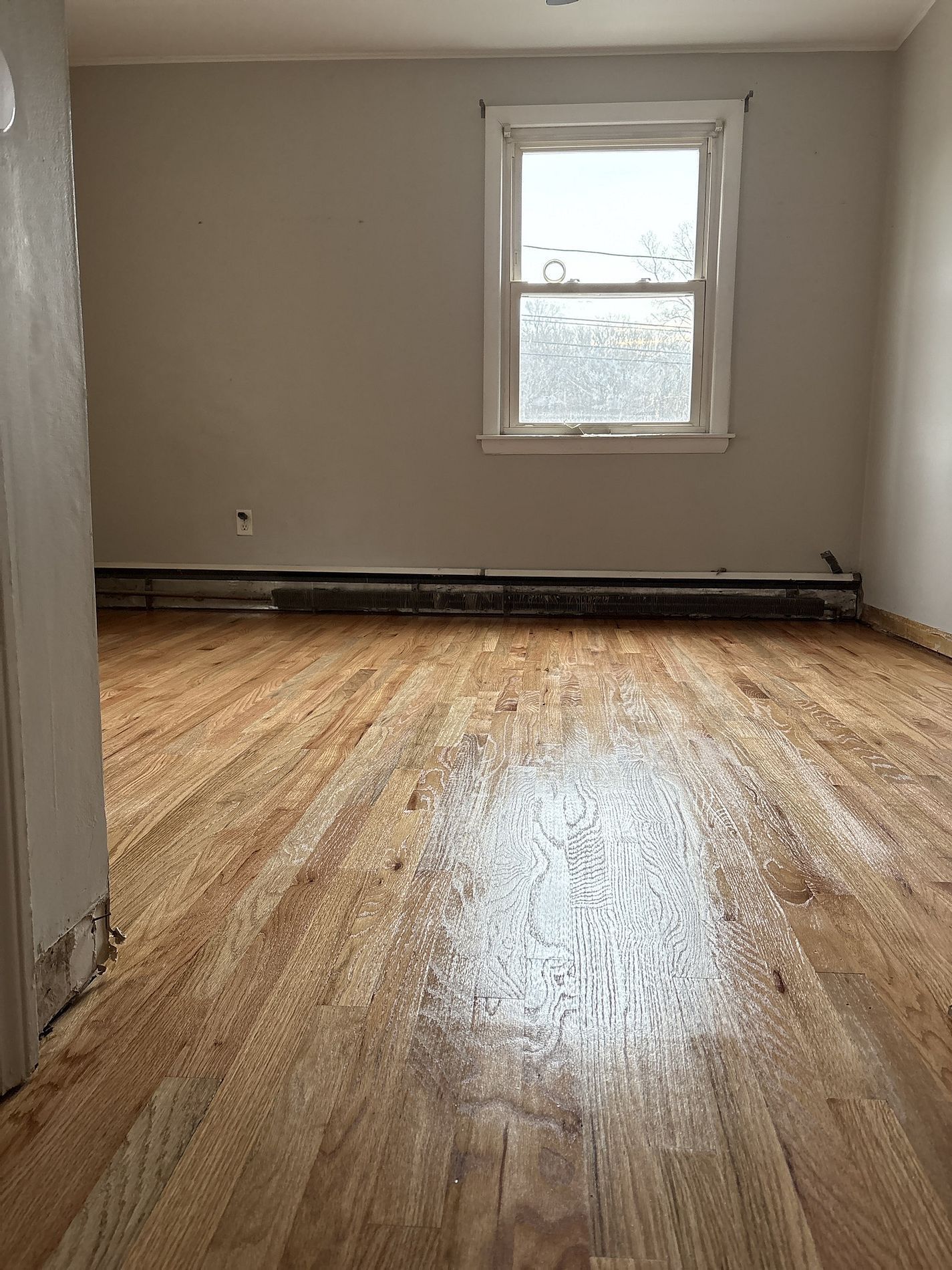Empty room with hardwood floors and a single window, showing wear and sunlit reflections.