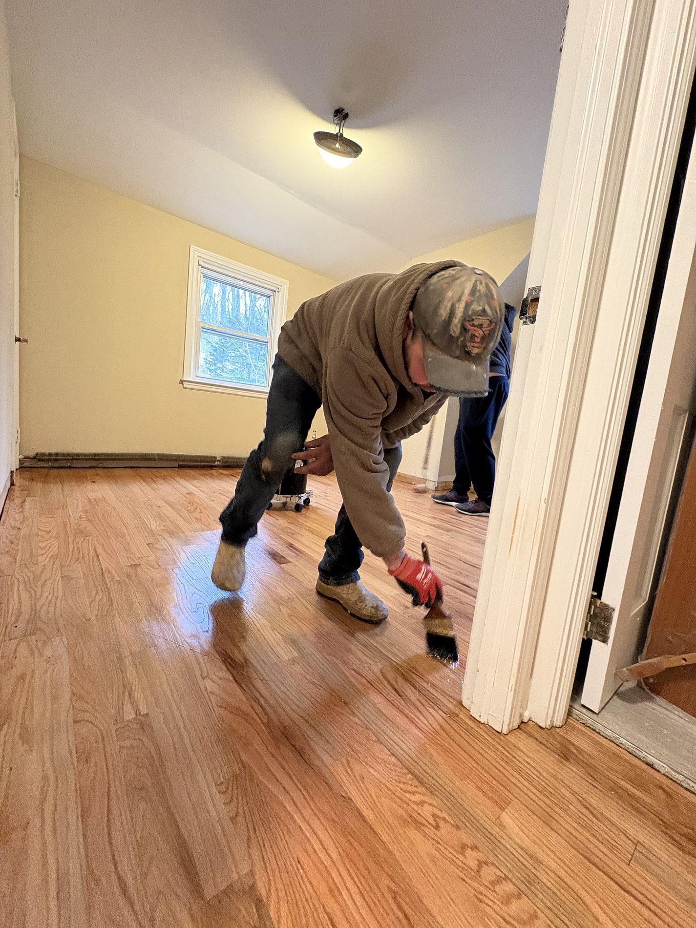 Worker cleaning a hardwood floor in an empty room with beige walls and a small window