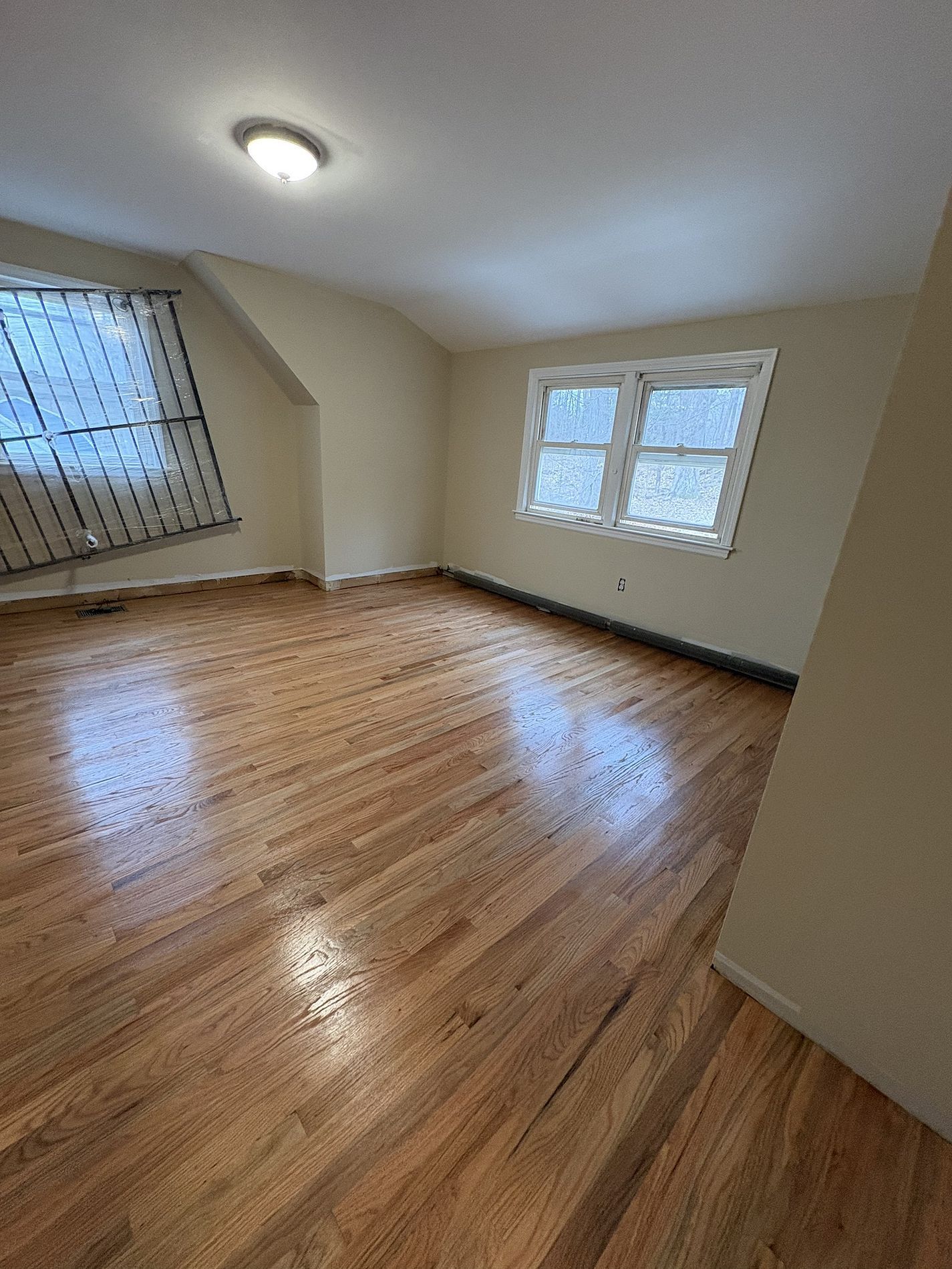 Empty room with hardwood floors, beige walls, and windows letting in daylight.