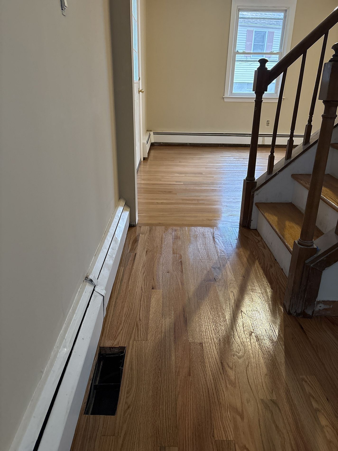 Narrow hallway with hardwood floor, staircase railing, white walls, and a window at the far end.