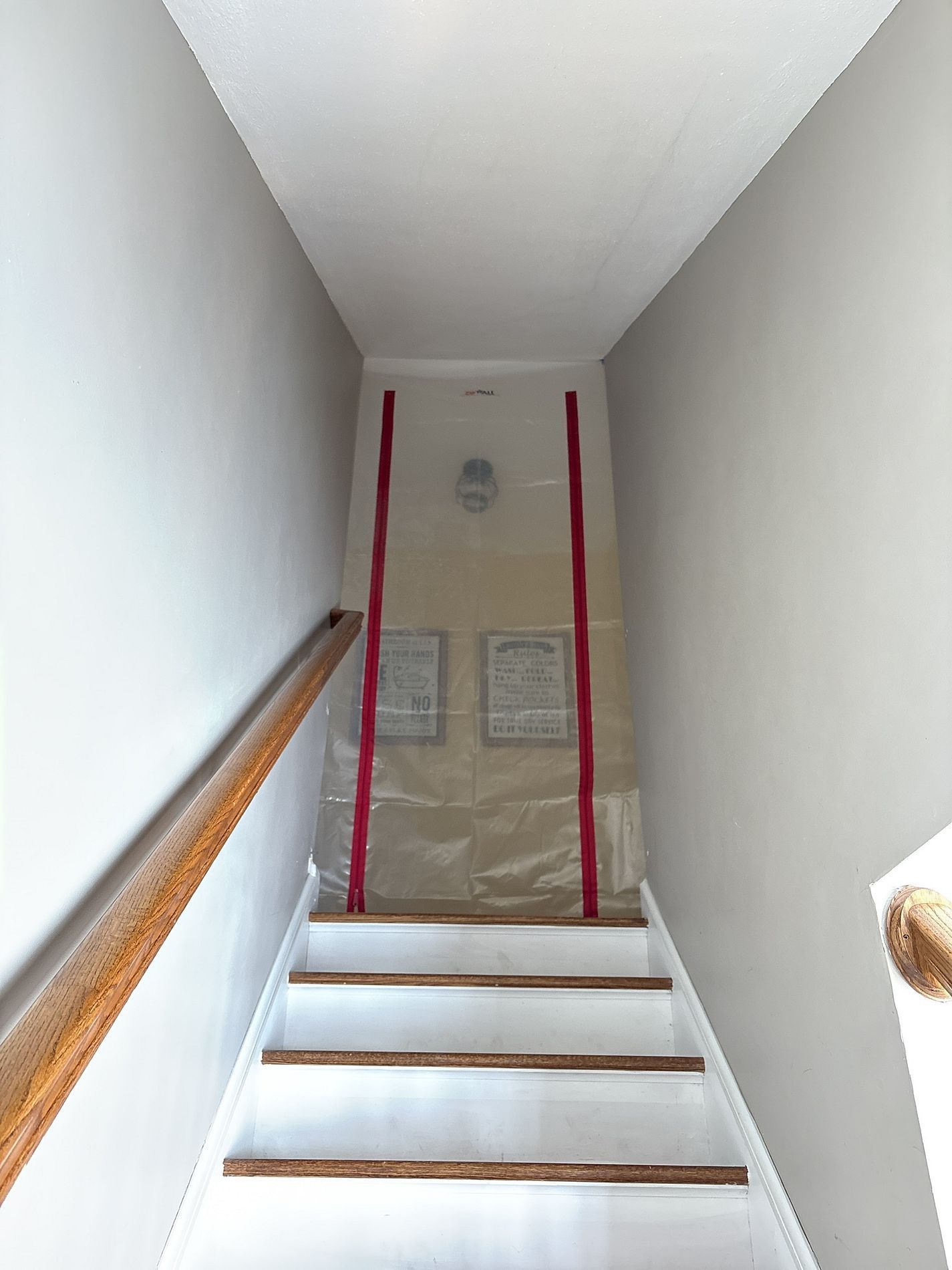 Narrow stairway with white walls, wooden handrail, and a plastic-covered doorway at the top