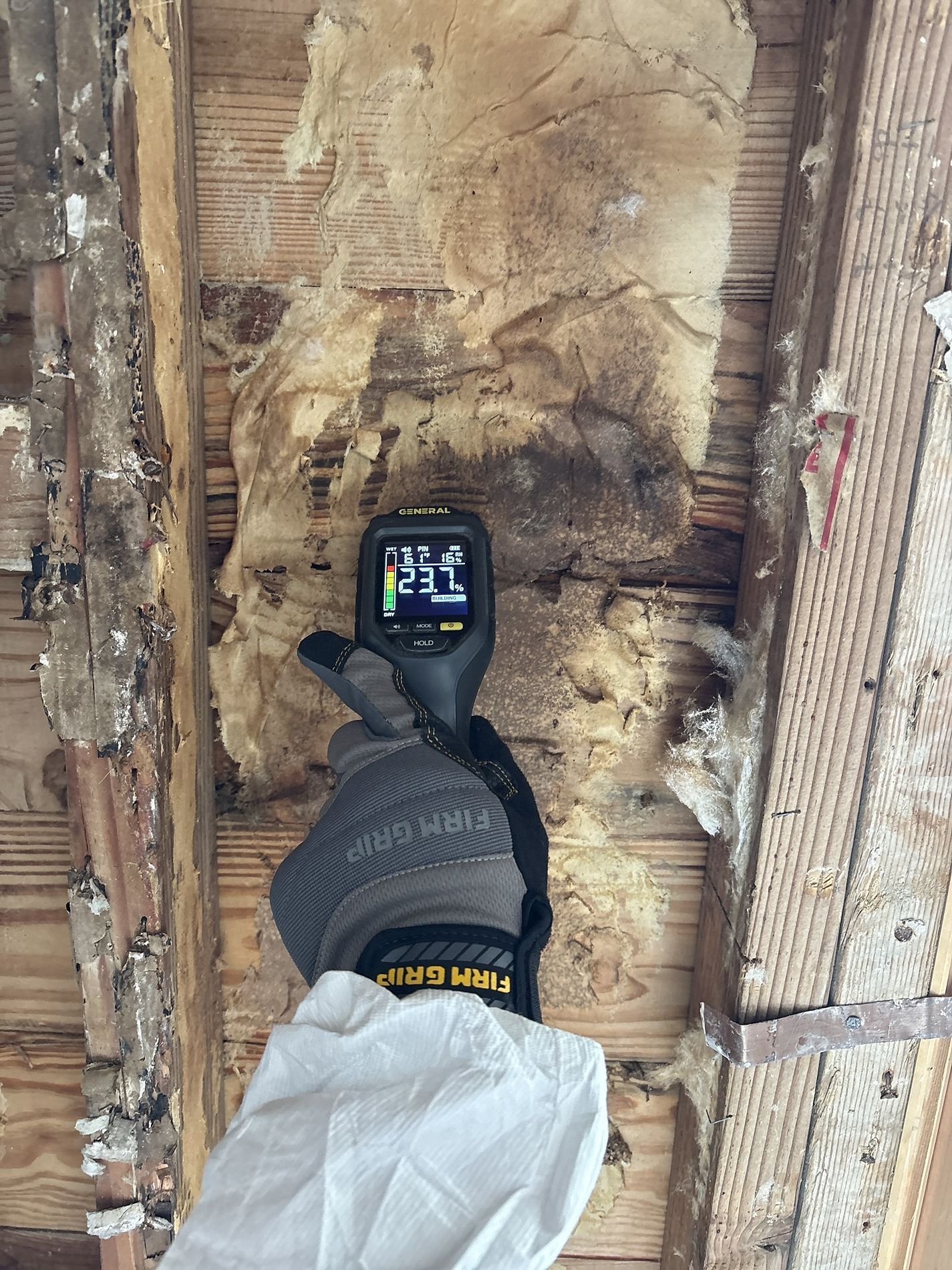 Gloved hand holding a thermal camera toward a damaged, exposed wall cavity with charred insulation.