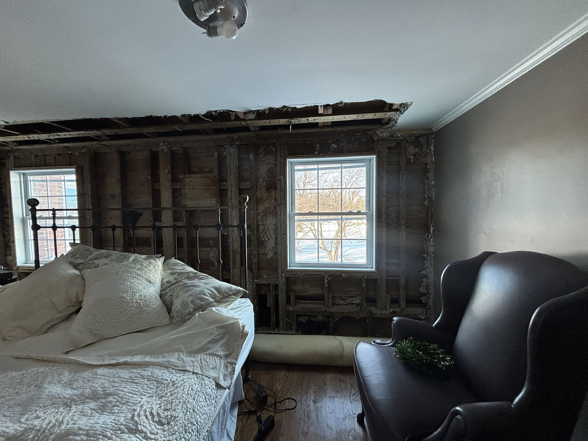 Partially renovated bedroom with exposed wall studs, a bed, a black chair, and a window with daylight.