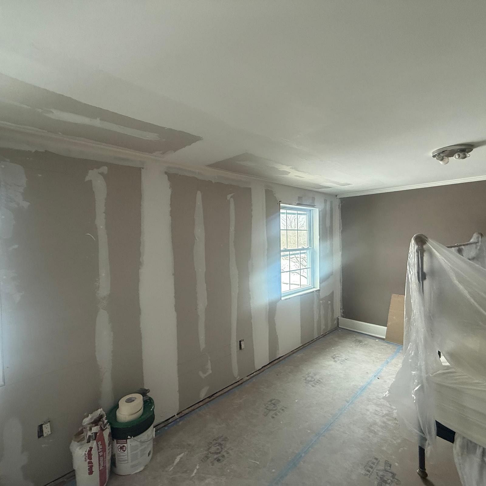 Partially renovated room with taped drywall, a window, and furniture covered in plastic.