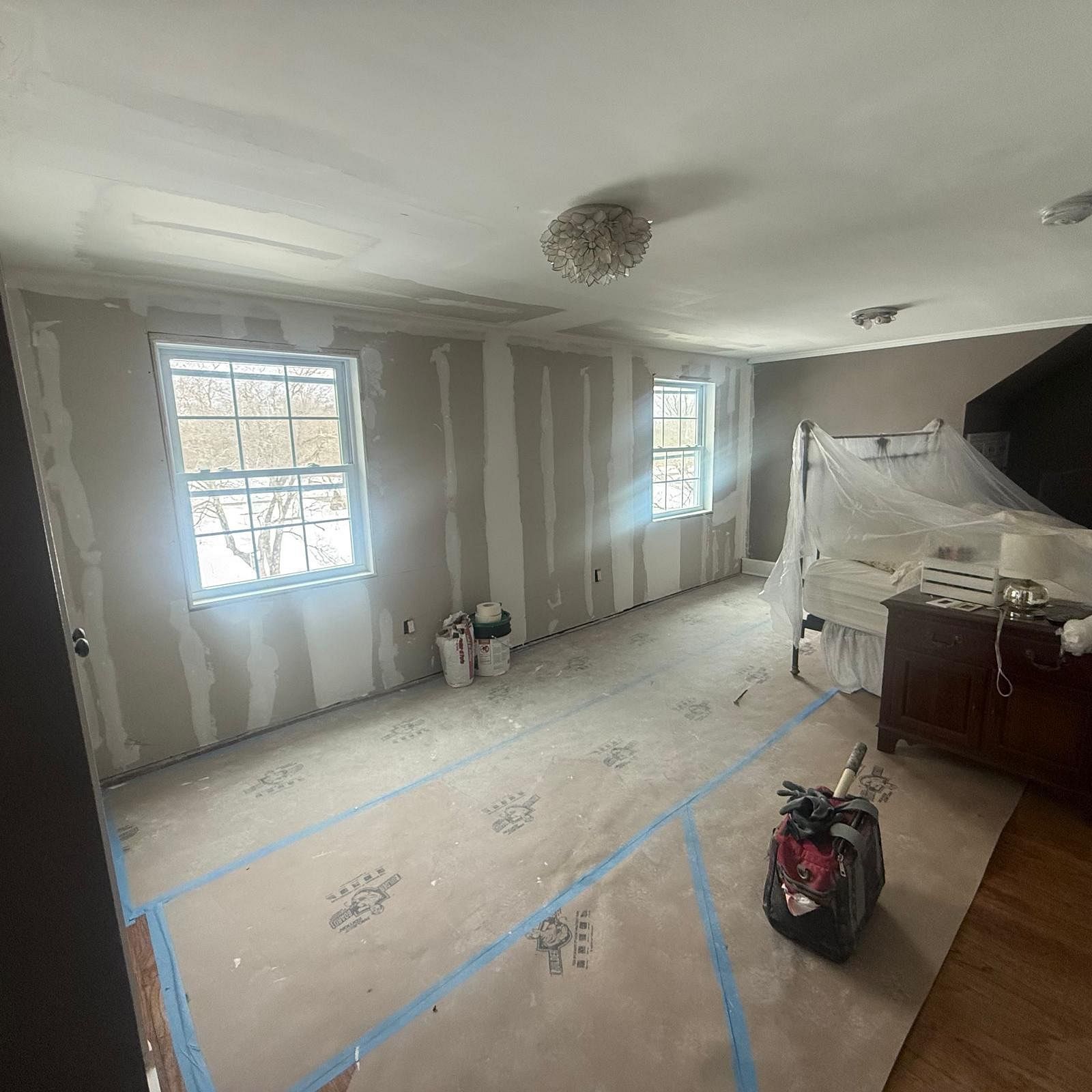Partially renovated room with taped floor, bare drywall, windows, covered furniture, and a ladder