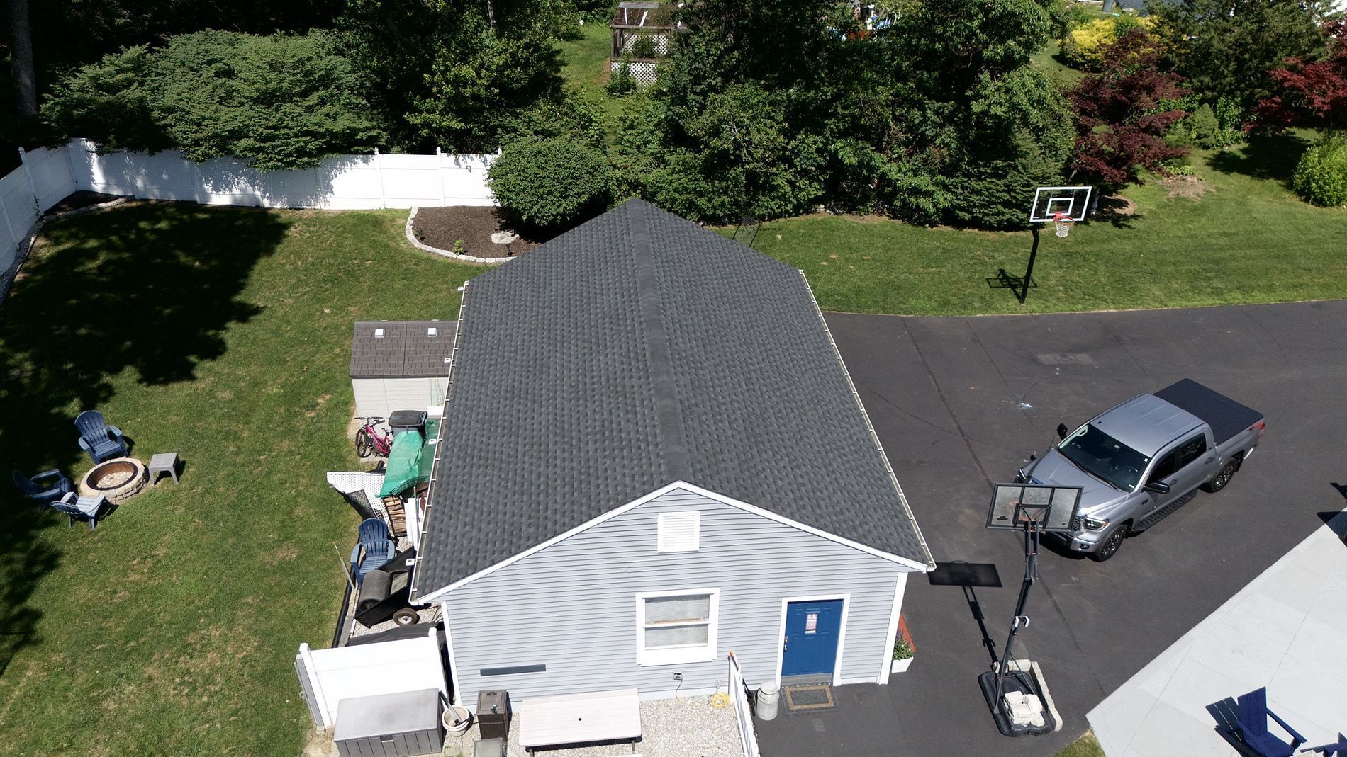 Aerial view of a small house with a gray roof, driveway, parked SUV, and a person near the side yard.