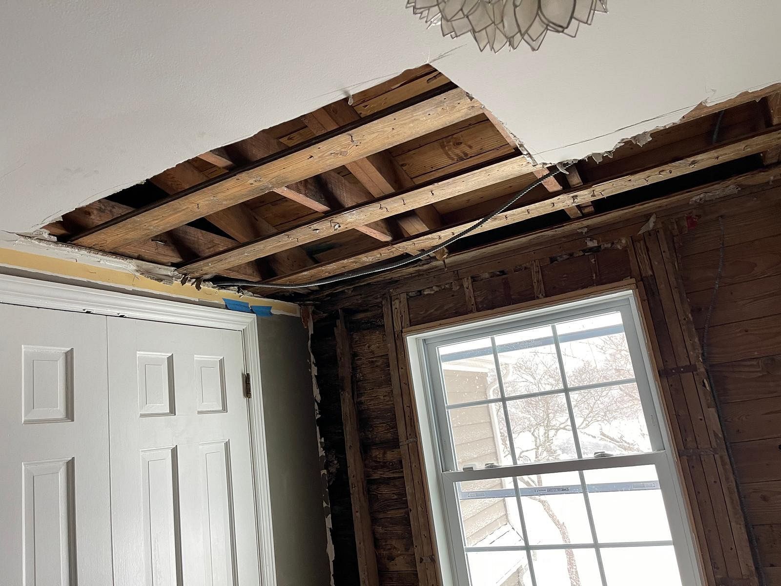 Damaged ceiling with exposed wood joists above a white closet door and window in a room.