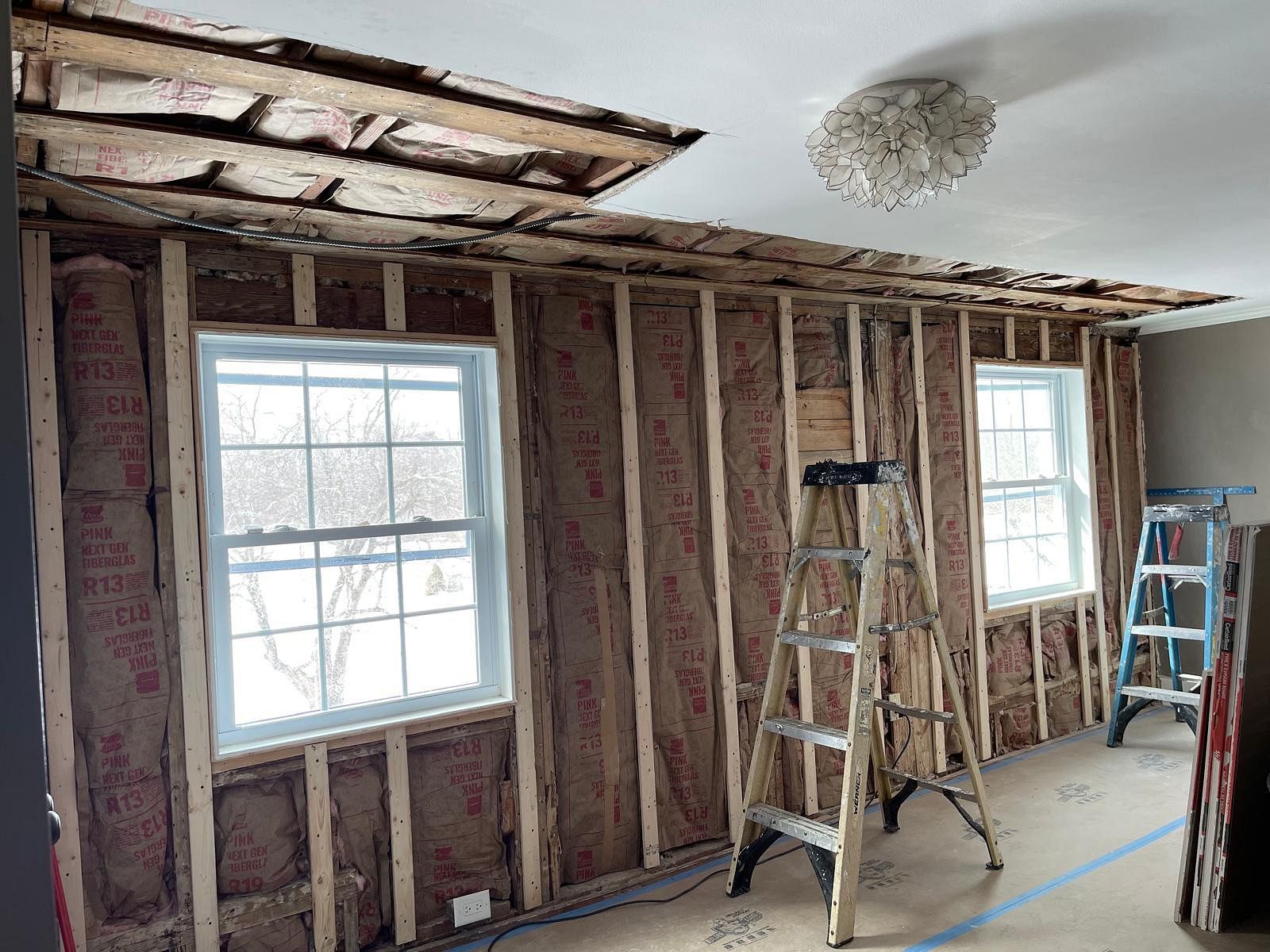 Partially demolished room with exposed studs, two windows, ladders, and a damaged ceiling