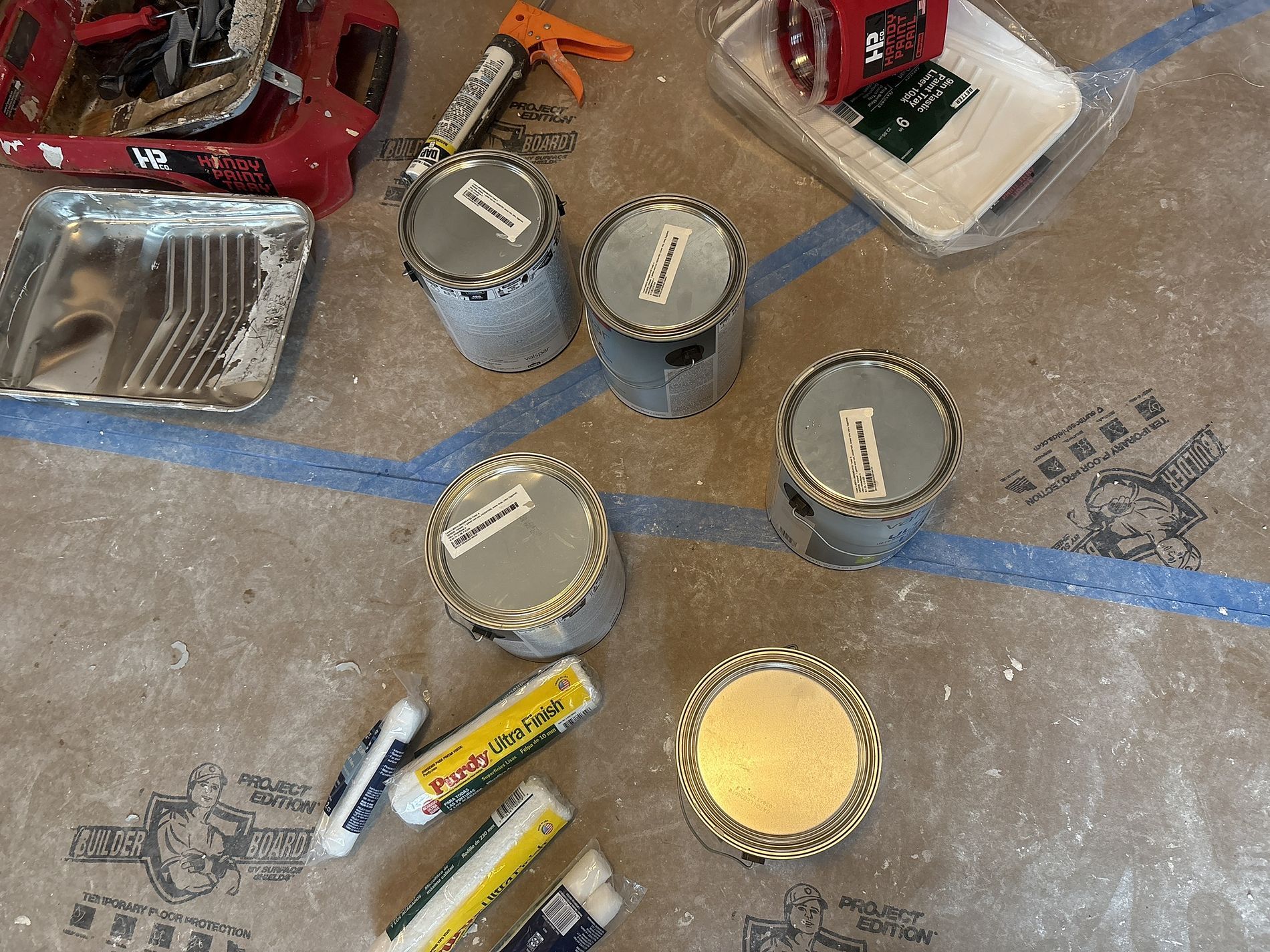 Paint cans, rollers, and supplies on a concrete floor marked with blue tape lines.