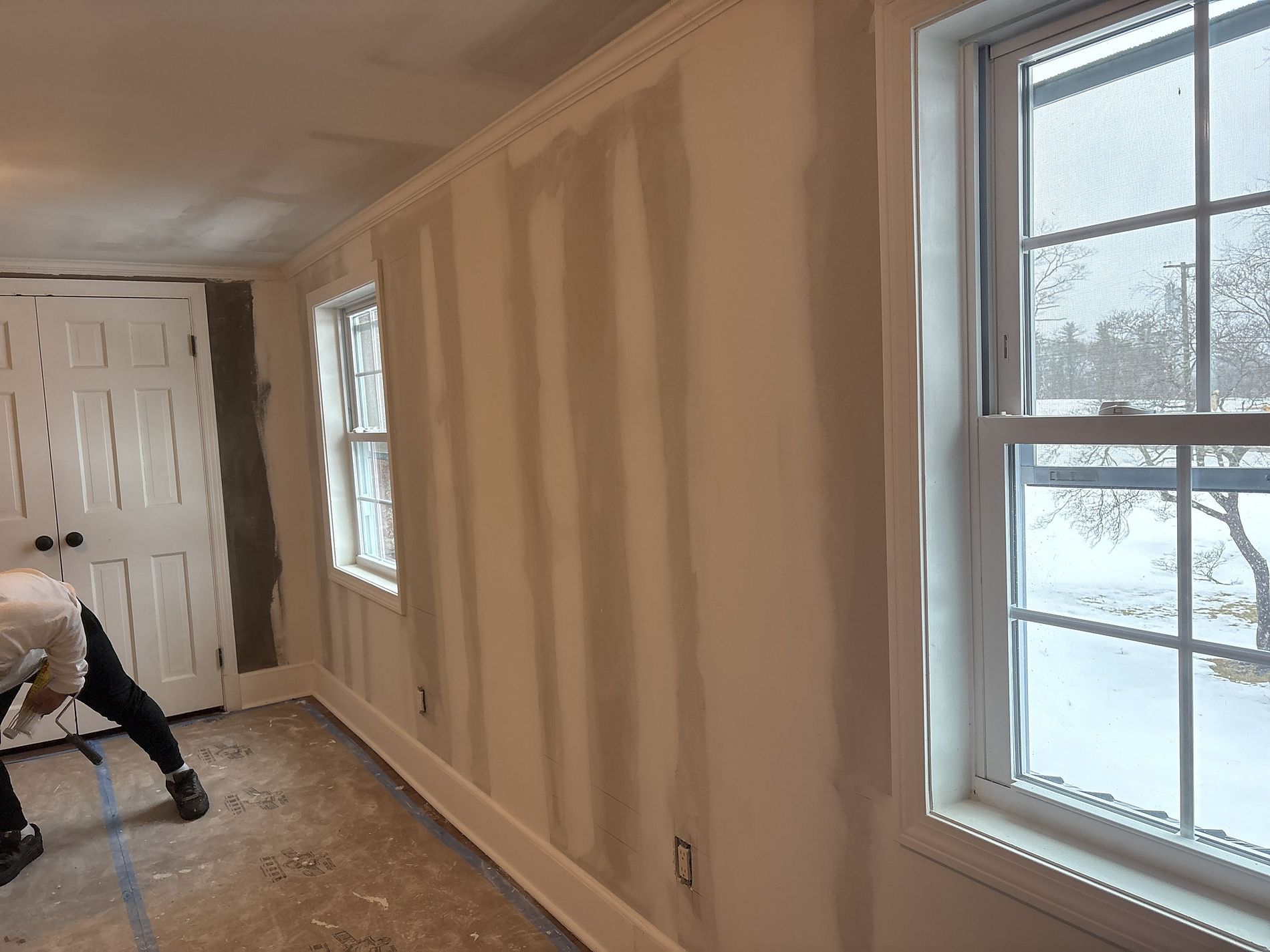 Interior room under renovation with unfinished drywall, windows, and a worker painting or sanding near the door.