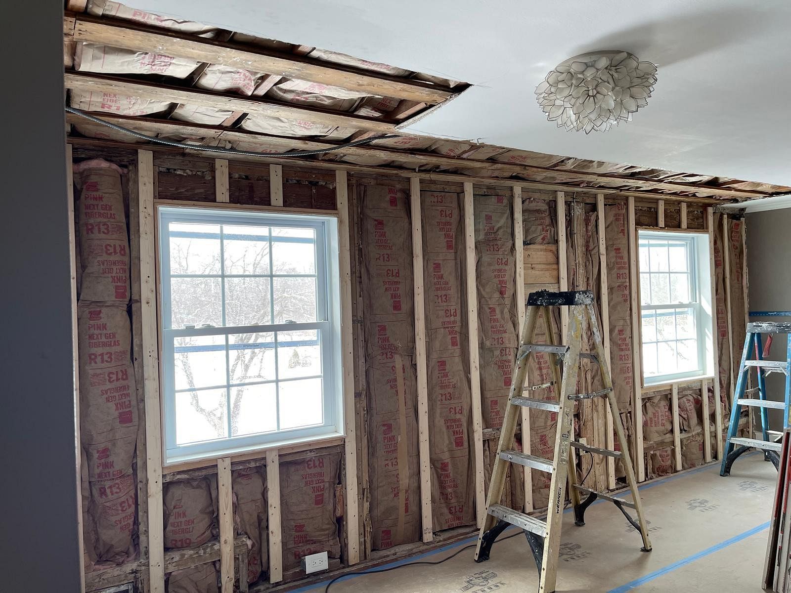 Interior renovation with exposed insulation, two windows, ladders, and debris on the floor