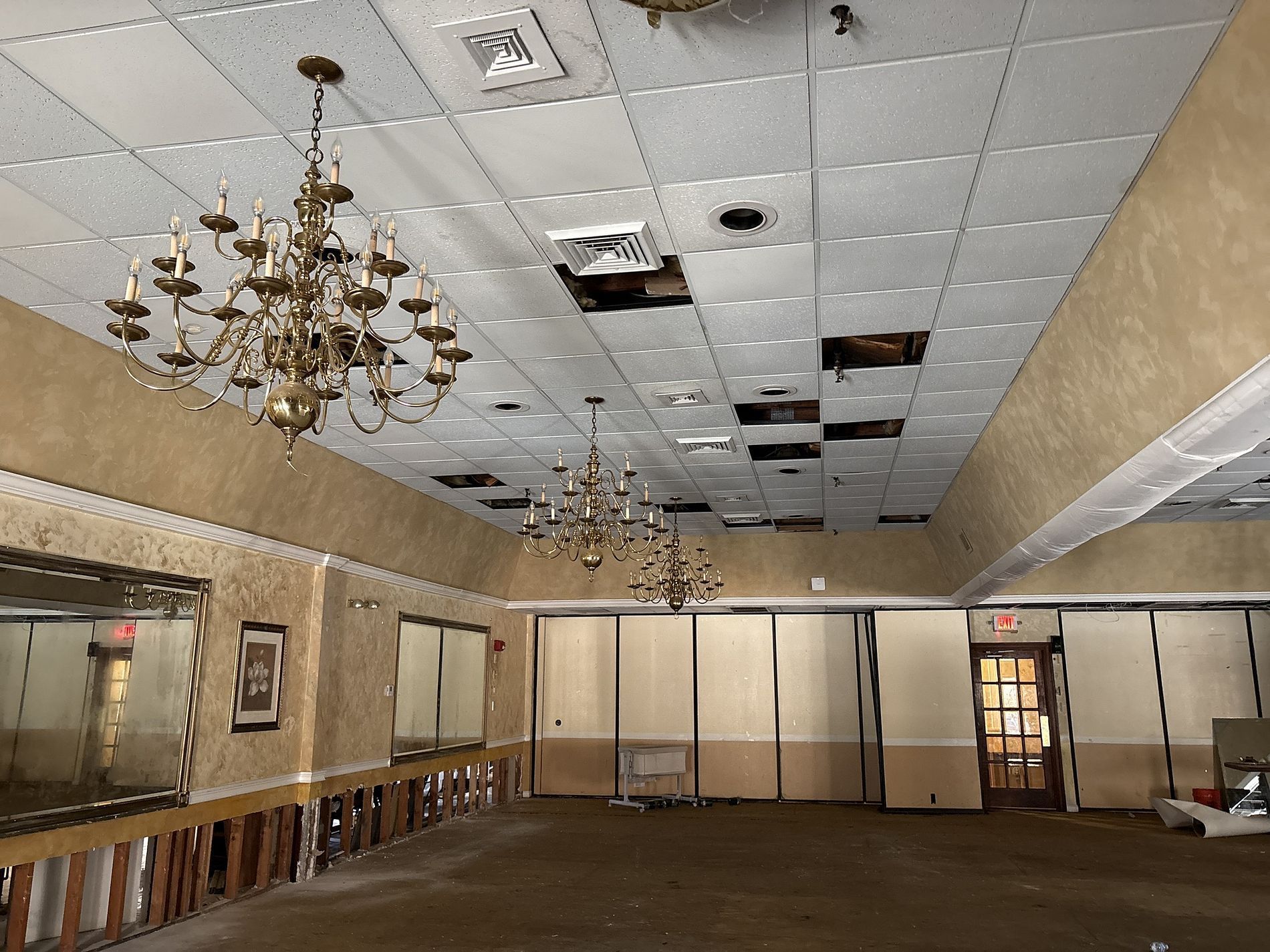 Damaged banquet hall ceiling with missing tiles, exposed holes, chandeliers, and beige walls