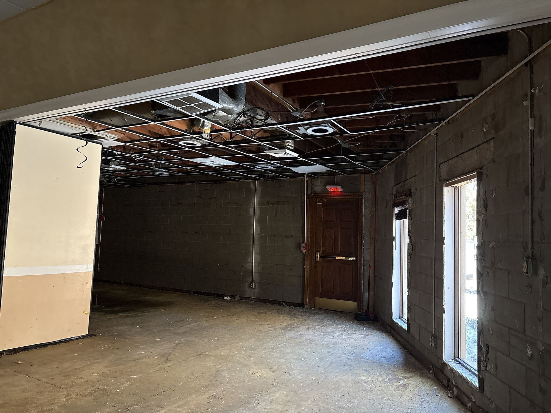 Damaged unfinished room with exposed ceiling beams, debris on floor, and tall windows on the right