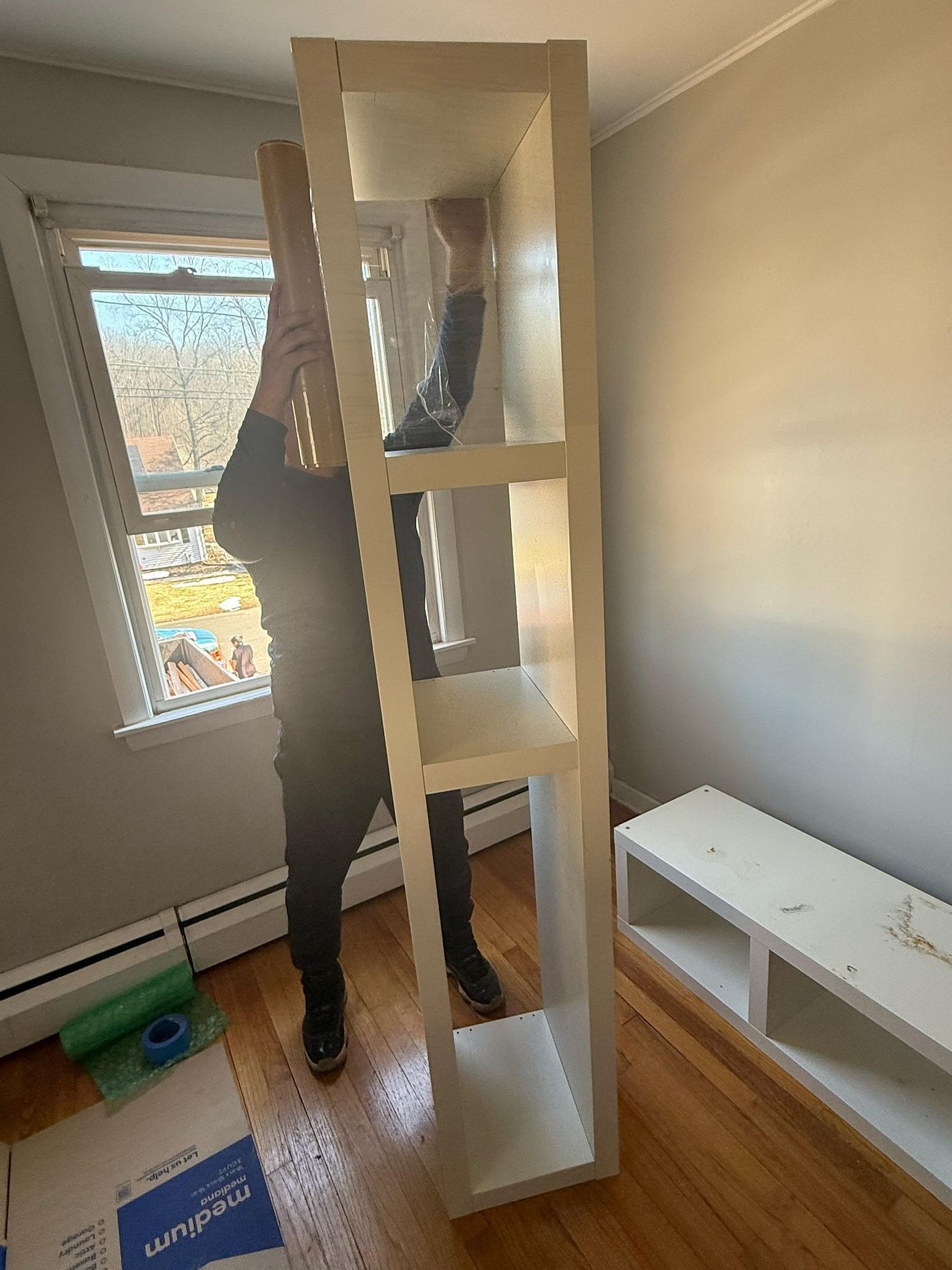 Person assembling a tall white shelving unit in a bright room beside a window.