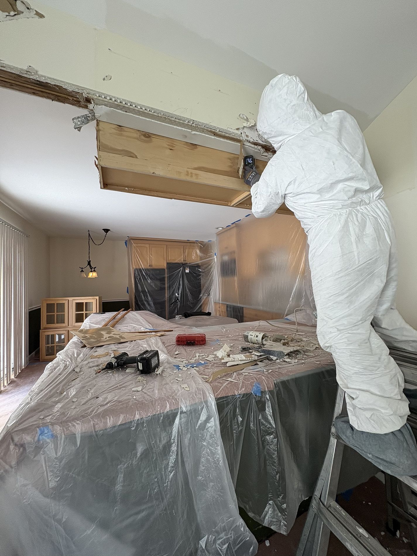 Worker in white protective suit demolishing a kitchen ceiling, with debris covered in plastic sheeting.