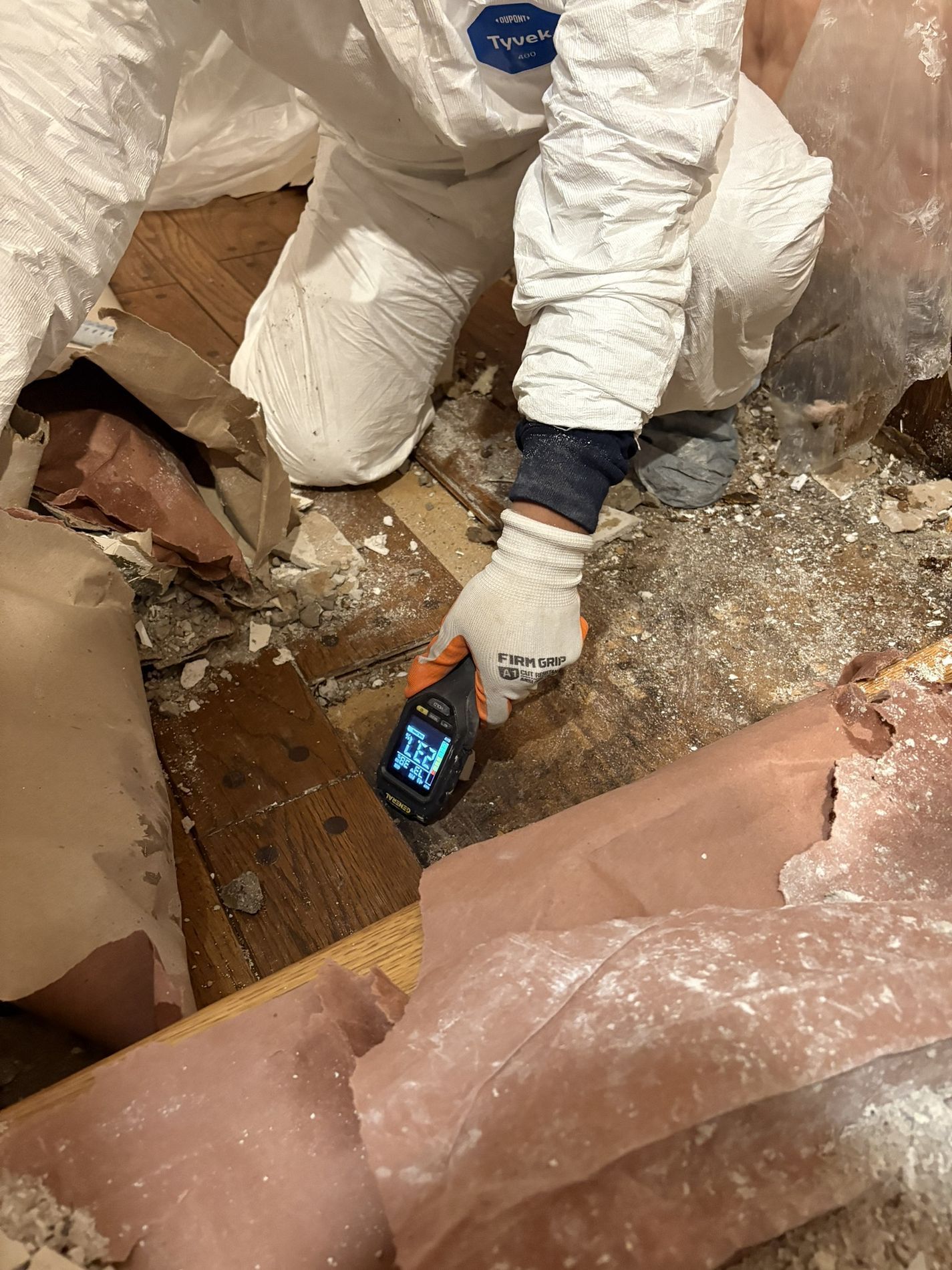 Worker in white protective suit using a handheld device to inspect debris in a damaged area.