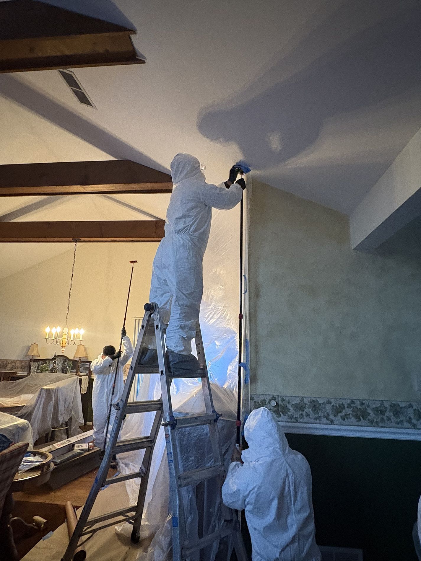 Two workers in white coveralls painting a ceiling in a living room, one on a ladder and one below.
