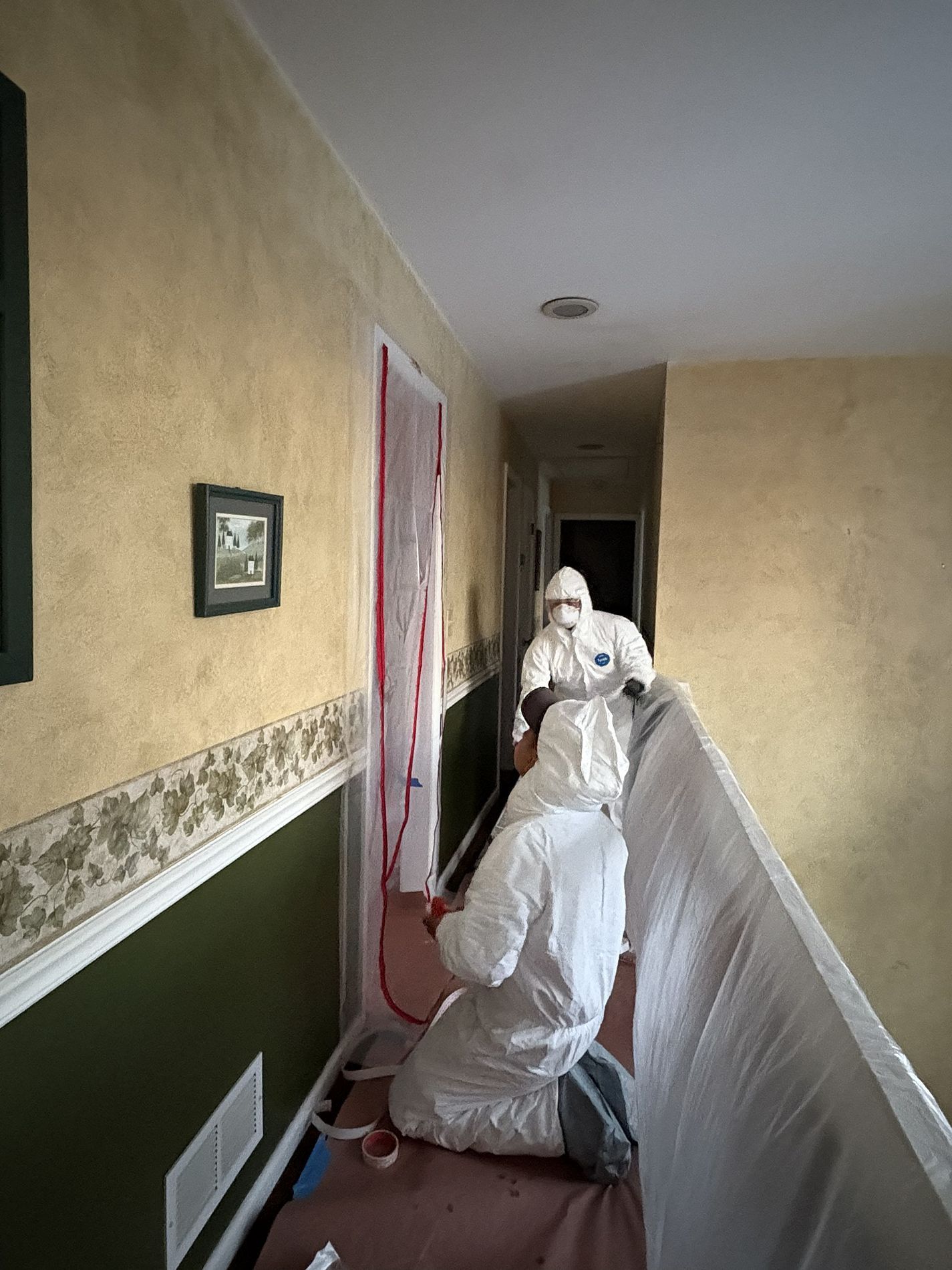 Two workers in white protective suits in a hallway, covering walls and floor with plastic sheeting.