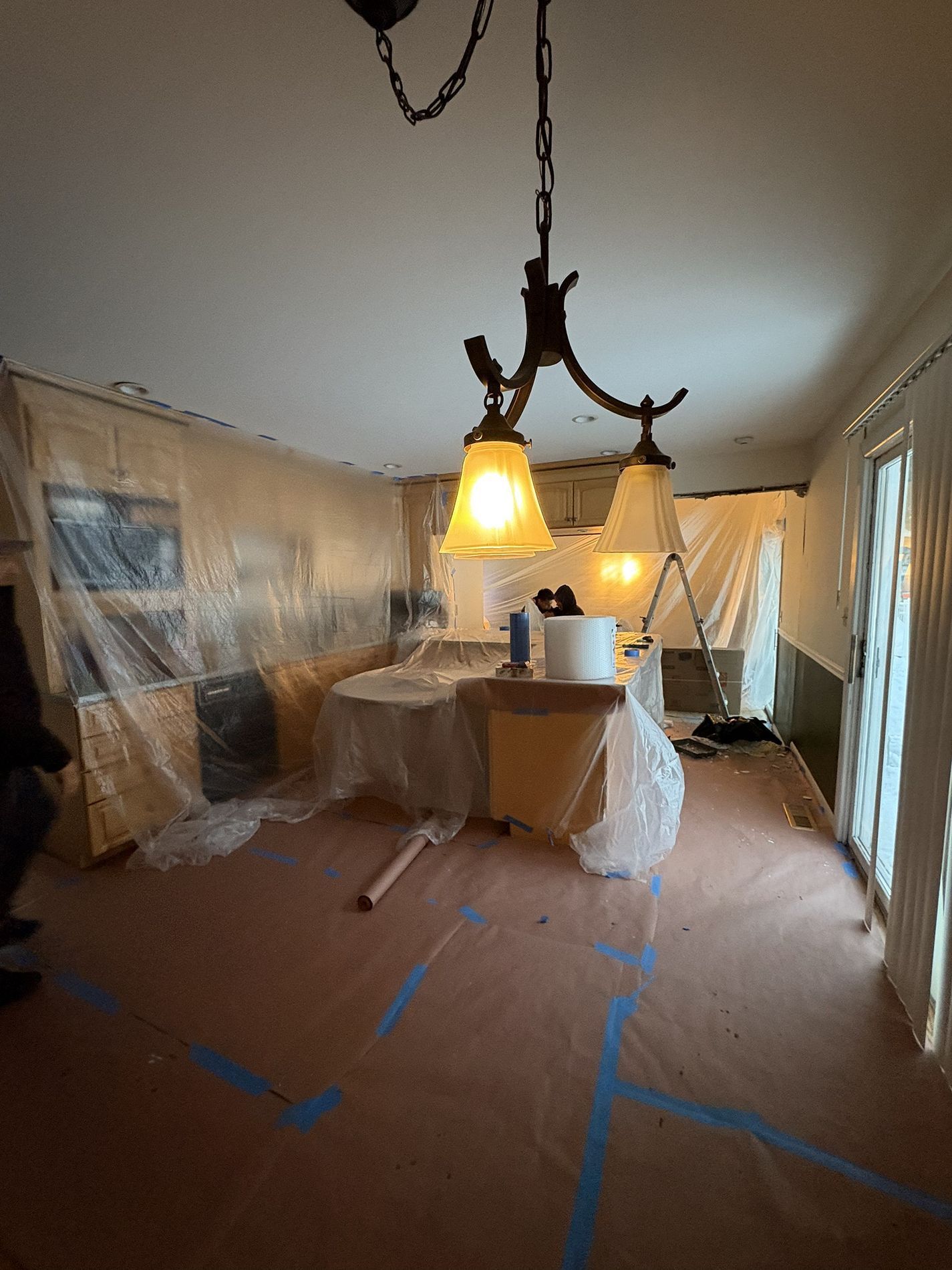 Dining room under renovation with covered furniture, plastic sheeting, and hanging light fixtures.