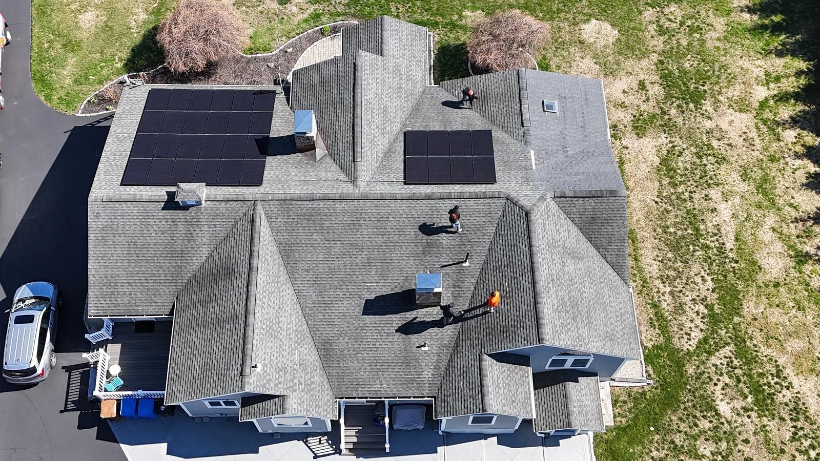 Aerial view of a gray roof with solar panels, skylights, and vents beside a driveway and parked vehicle.