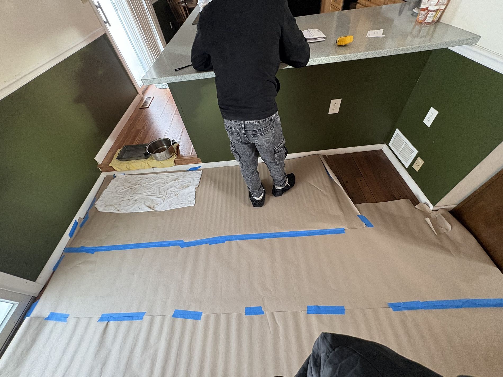 Person standing in a taped-off kitchen area with cardboard floor protection and cabinets along the wall