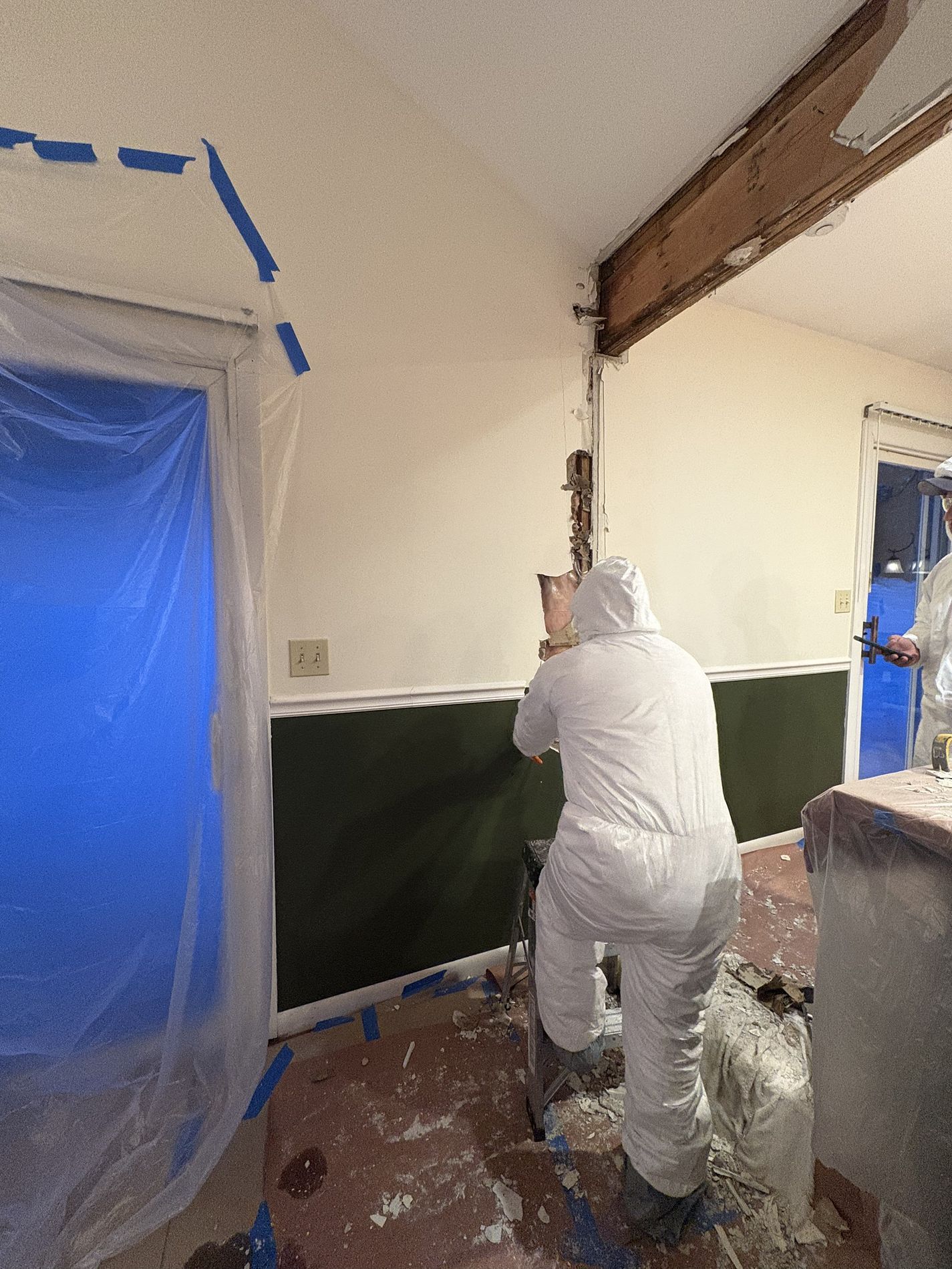 Person in white coveralls painting a wall inside a room with blue-taped door and plastic sheeting