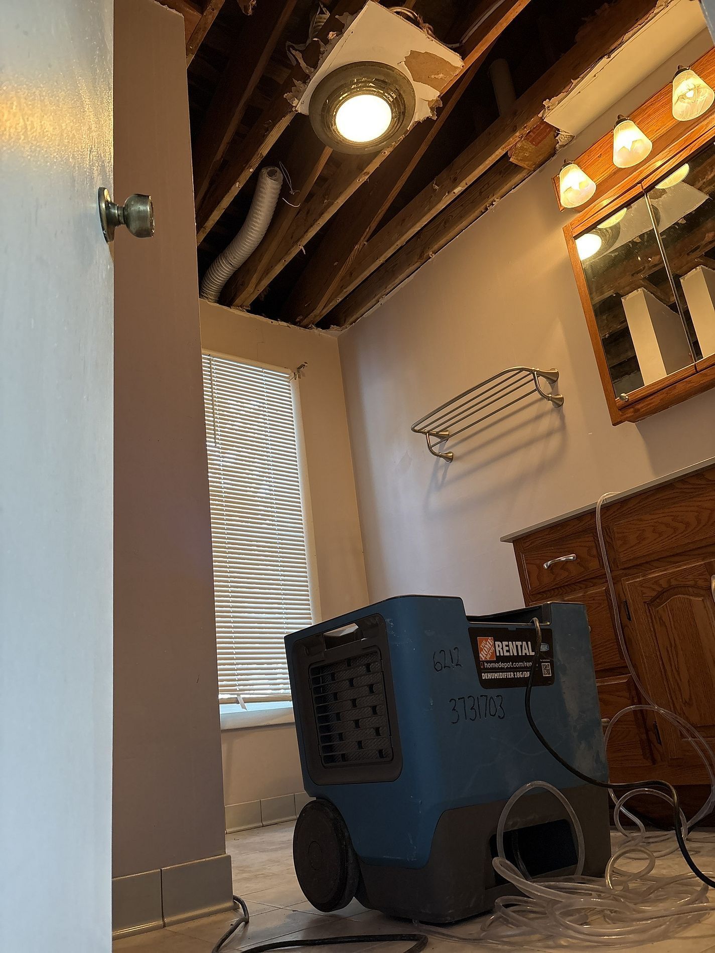 Bathroom corner with a blue portable dehumidifier near a vanity, window, and hanging wall rack.