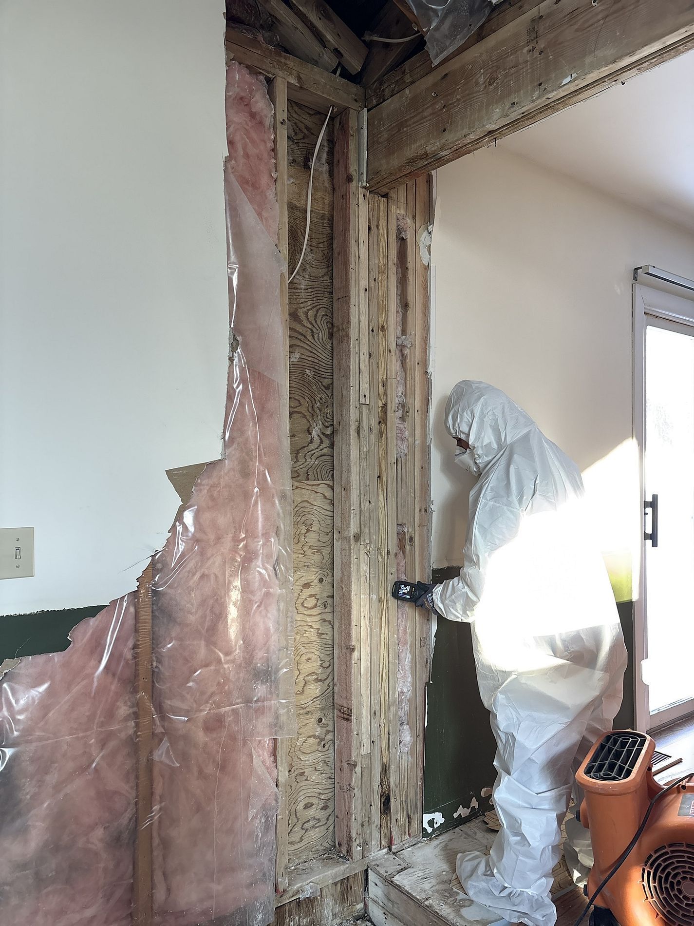 Worker in white protective suit inspects exposed wall framing during demolition inside a damaged room.