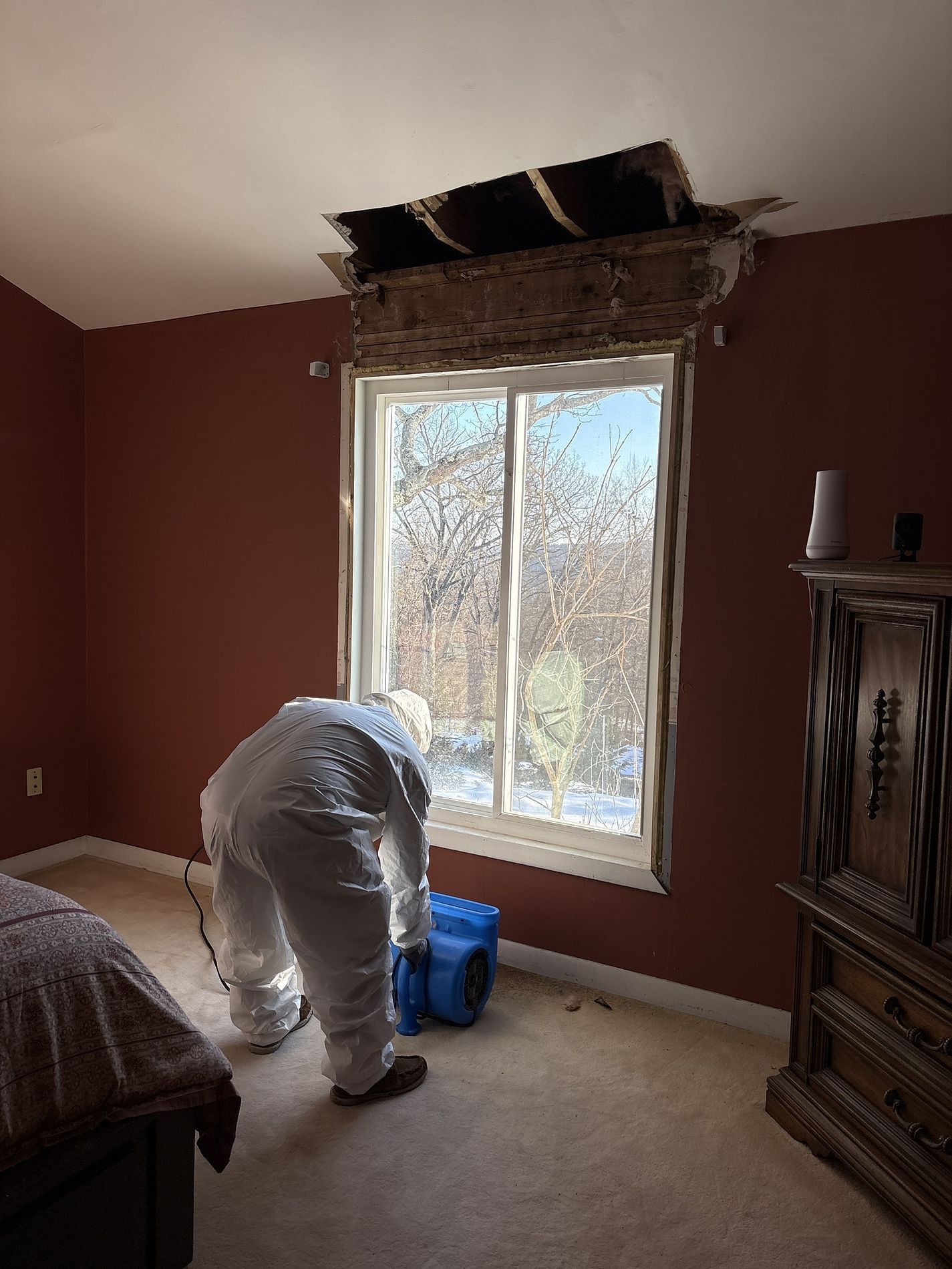 Worker in protective suit near a window in a room with ceiling damage and repair materials.