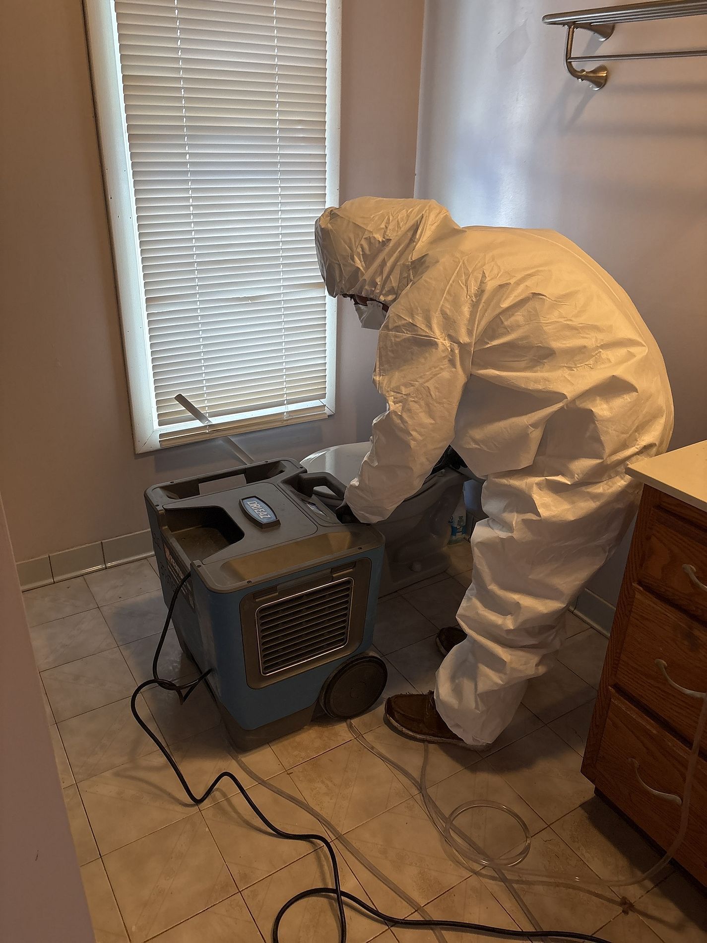 Person in white protective suit inspecting a gray machine in a tiled room with blinds.