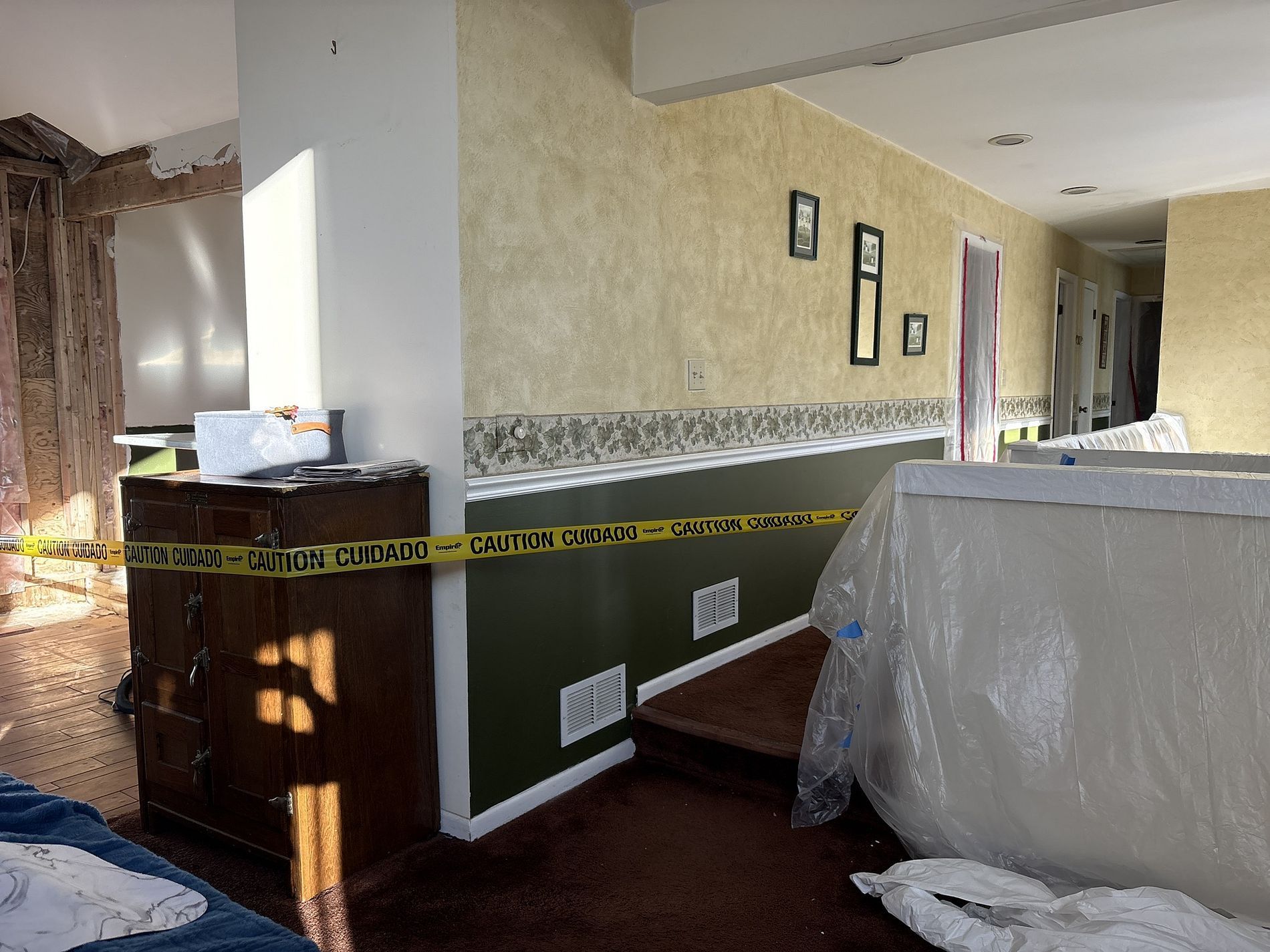 Caution tape across a hallway with exposed wall damage and covered furniture in a damaged room