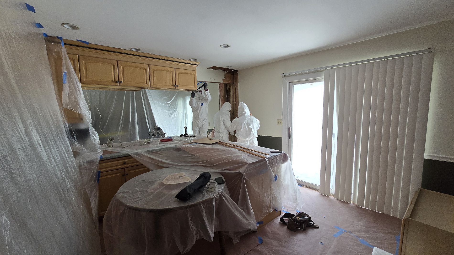 Living room covered in plastic for renovation, with sliding glass doors and a person standing by the counter.
