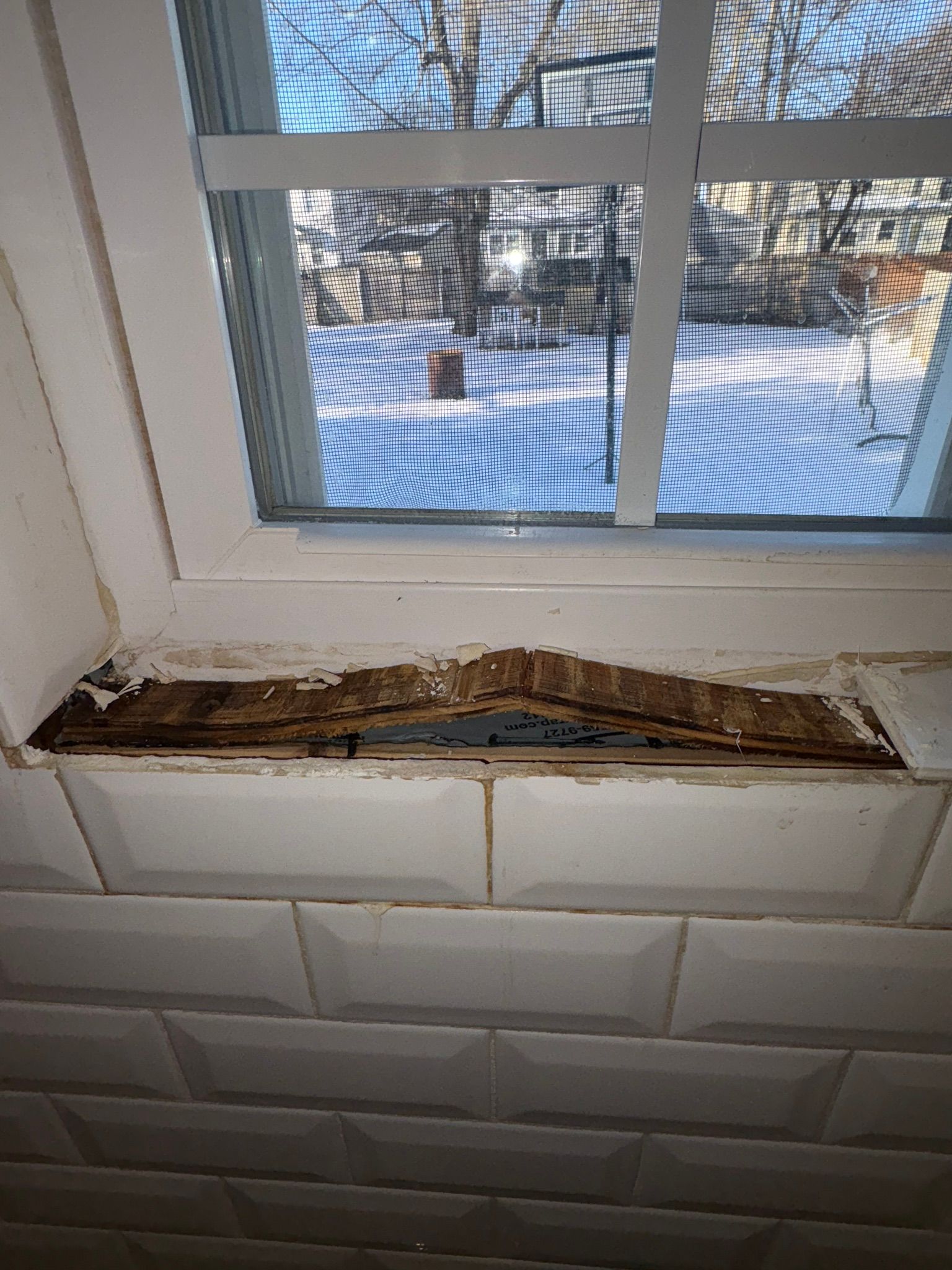 Damaged window sill with peeling wood beneath a white window, showing snow outside.