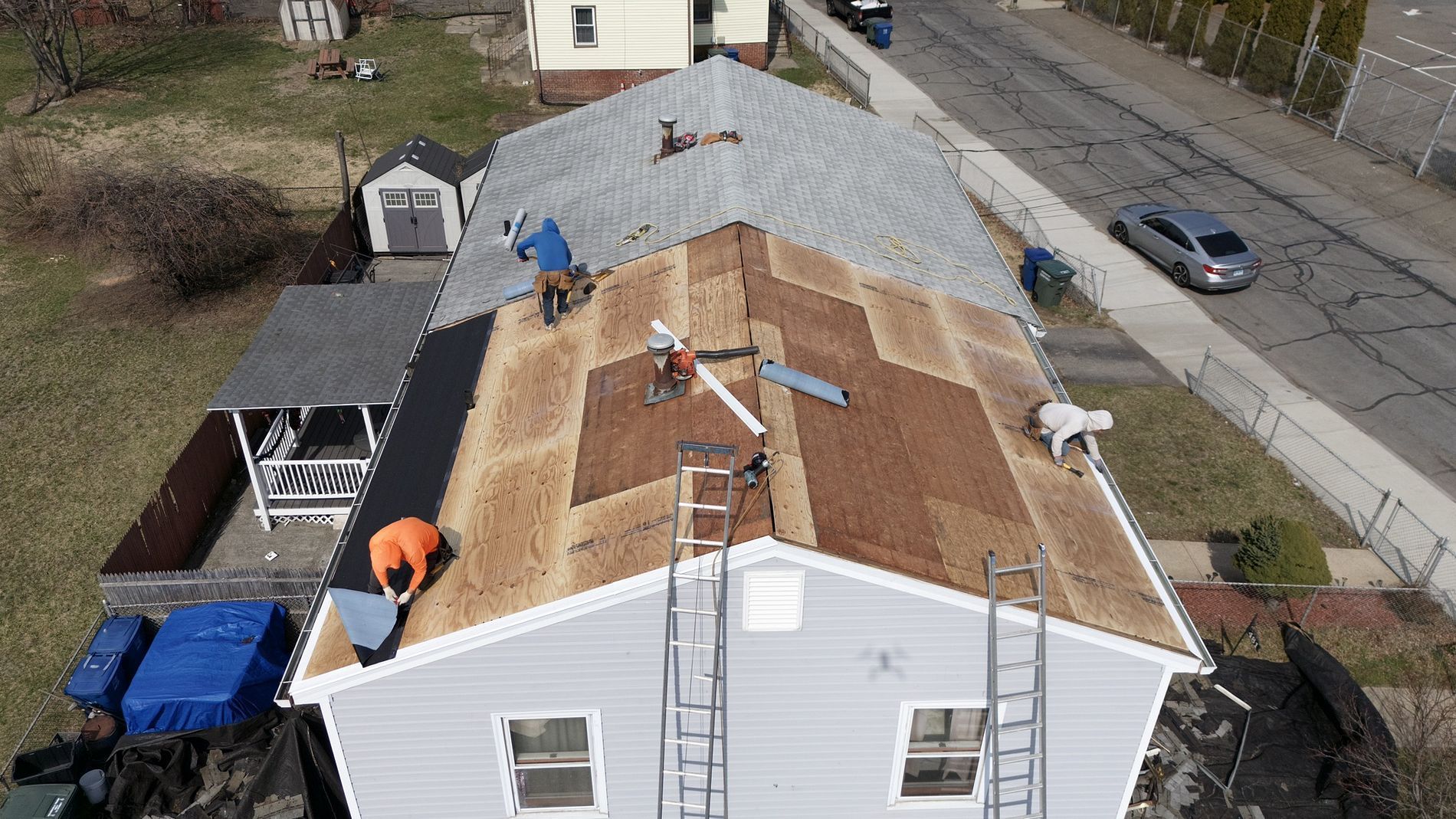 Workers installing a new roof on a white house, with exposed plywood and gray shingles in progress.