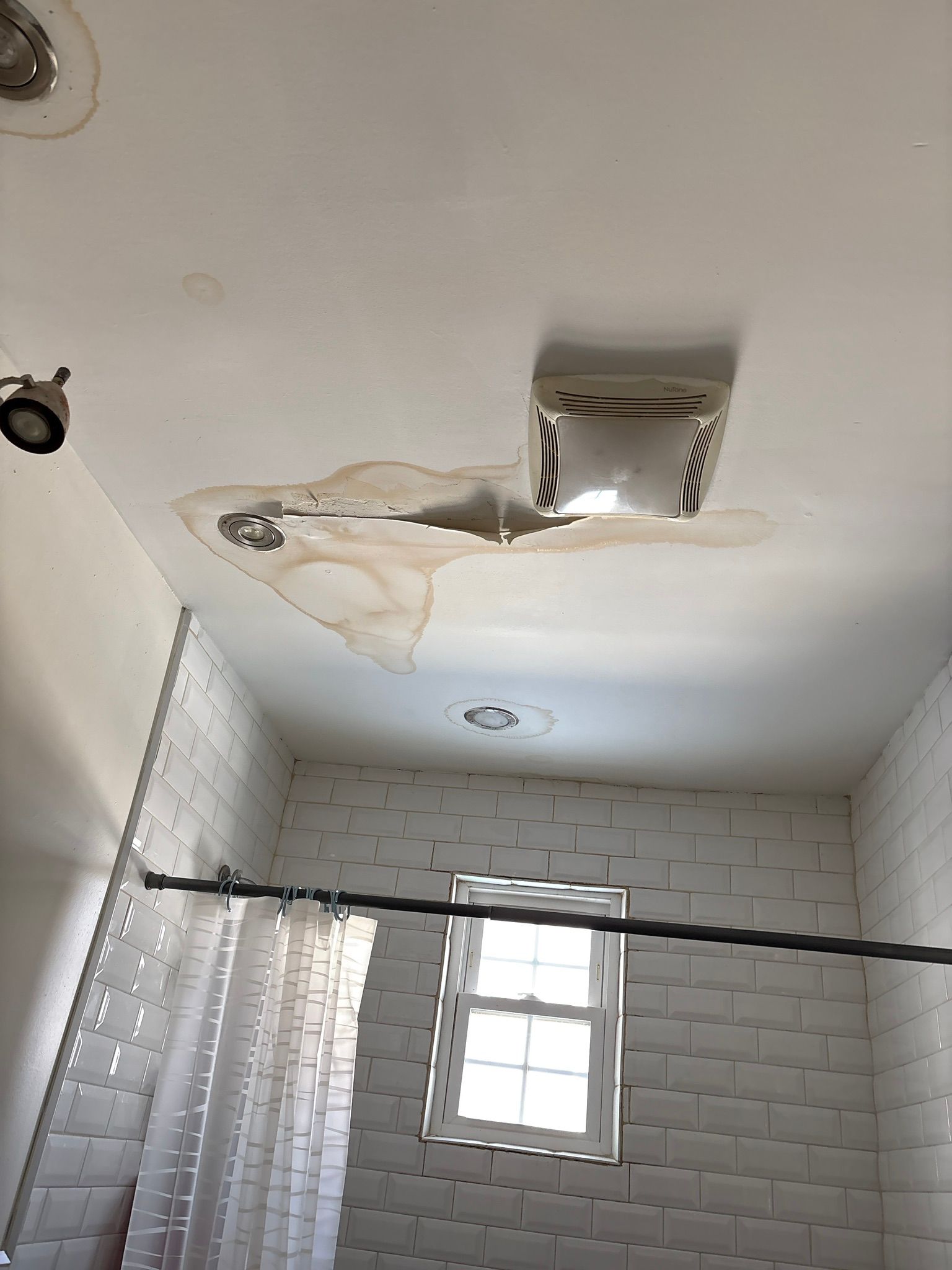 Bathroom ceiling with water damage, peeling paint, and a small window above white tiled walls