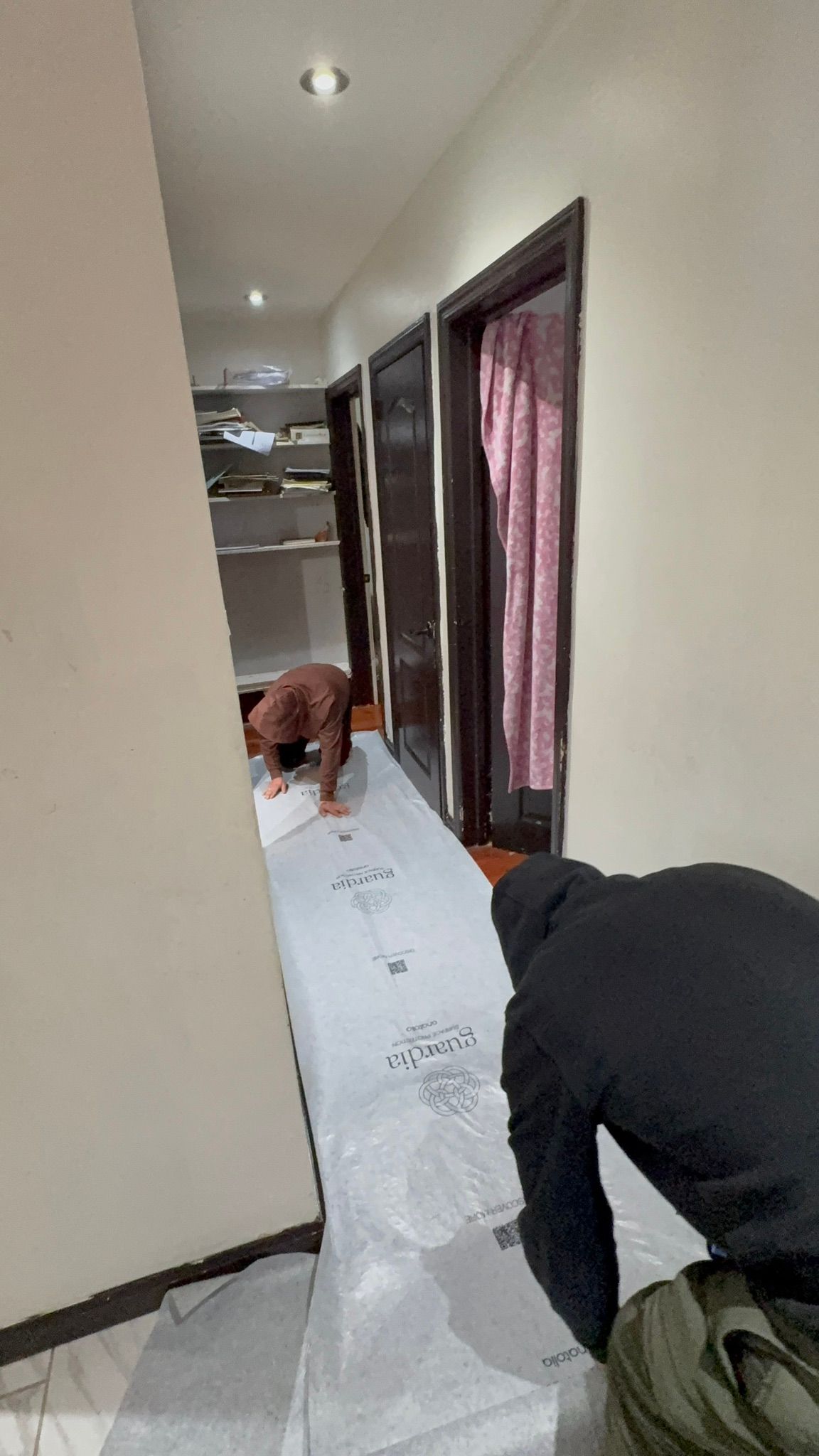 Two workers laying a white floor covering in a narrow hallway with doors along the side.